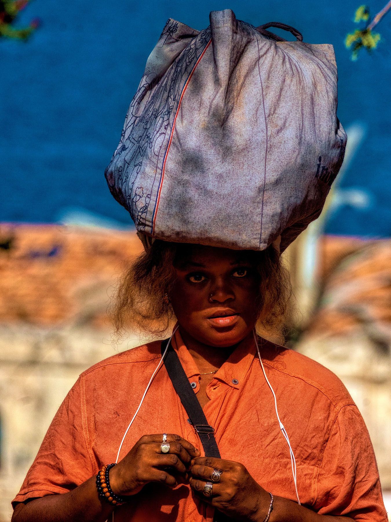 A person in an orange shirt carries a large bag on their head and looks at the viewer.