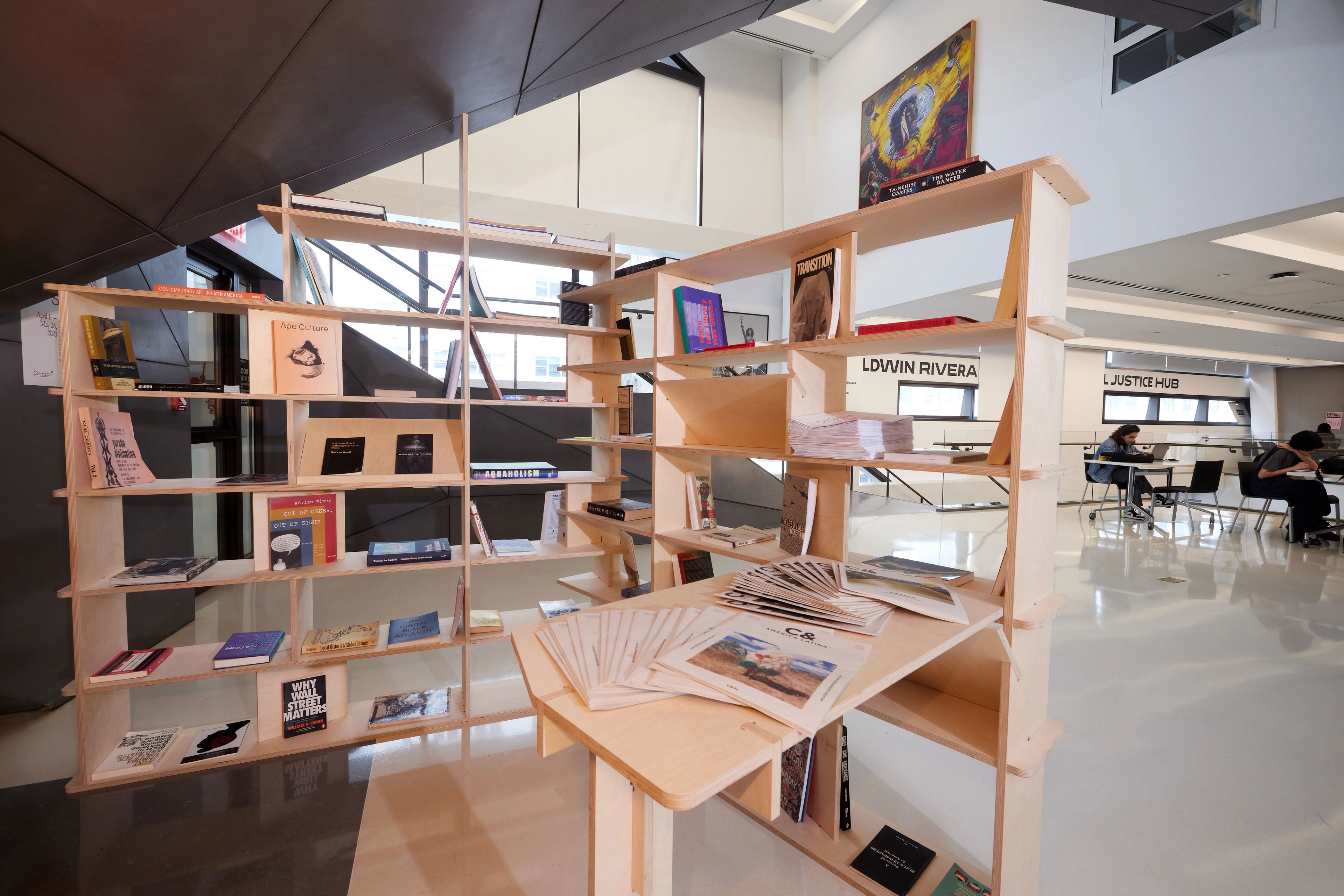 A modular light wood bookshelf stocked with books and magazines in a bright, modern indoor space with people working in the background.