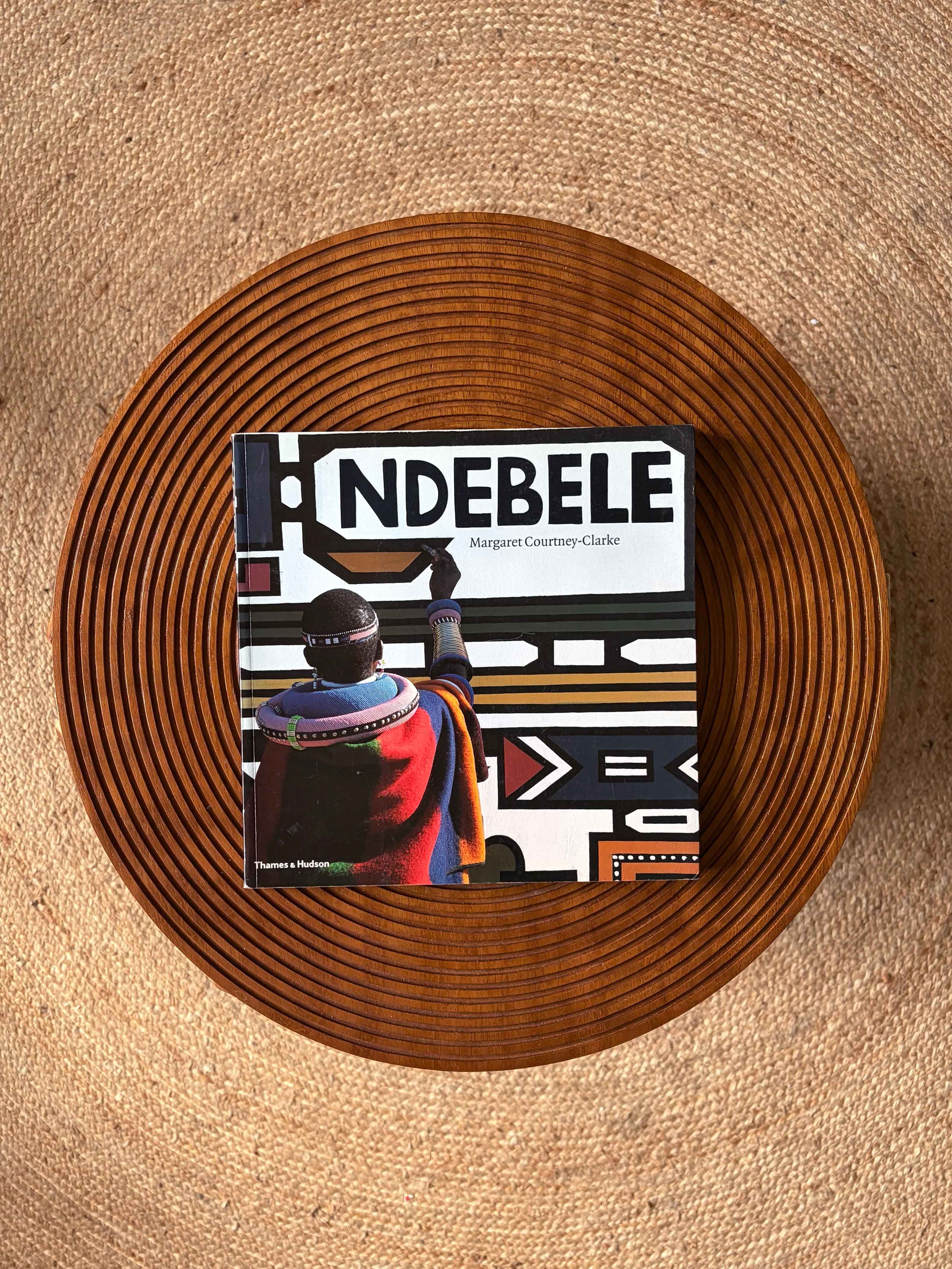 The book "NDEBELE" by Margaret Courtney-Clarke, showing a Ndebele person painting geometric art, rests on a round wooden tray atop a textured rug.
