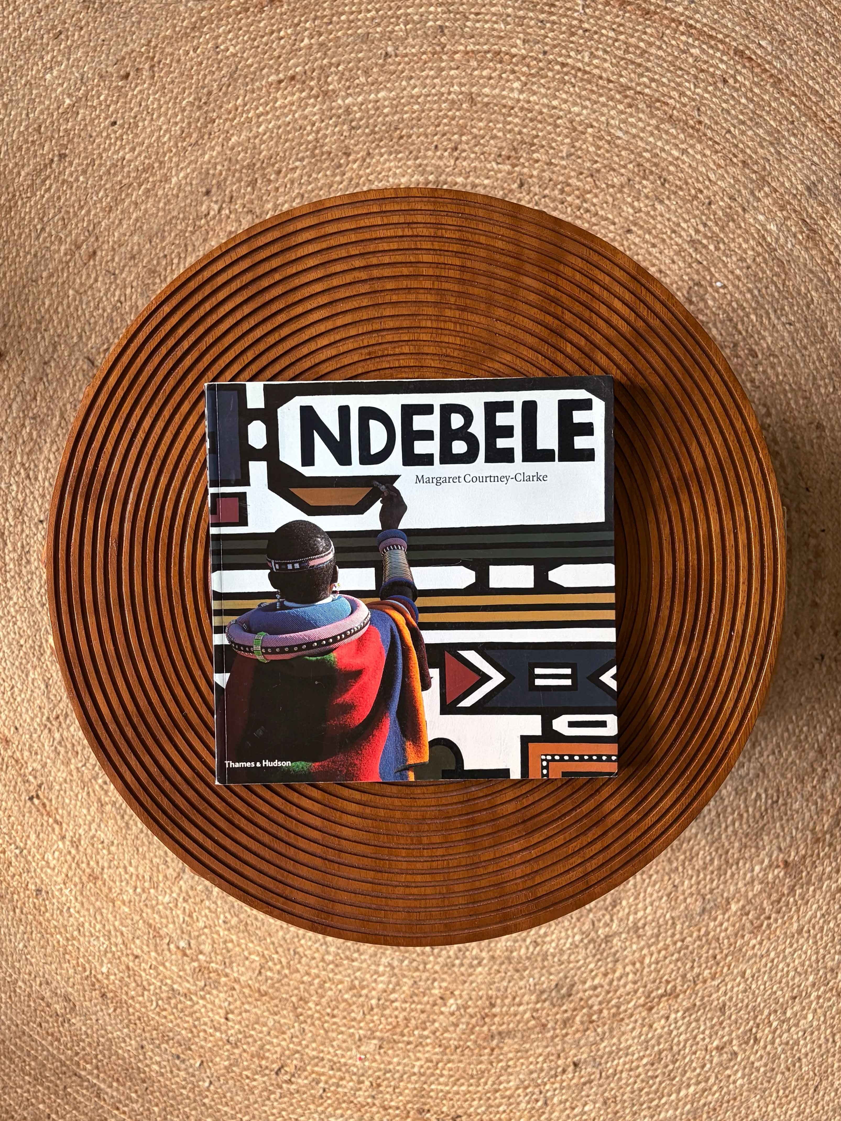 The book "NDEBELE" by Margaret Courtney-Clarke, showing a Ndebele person painting geometric art, rests on a round wooden tray atop a textured rug.