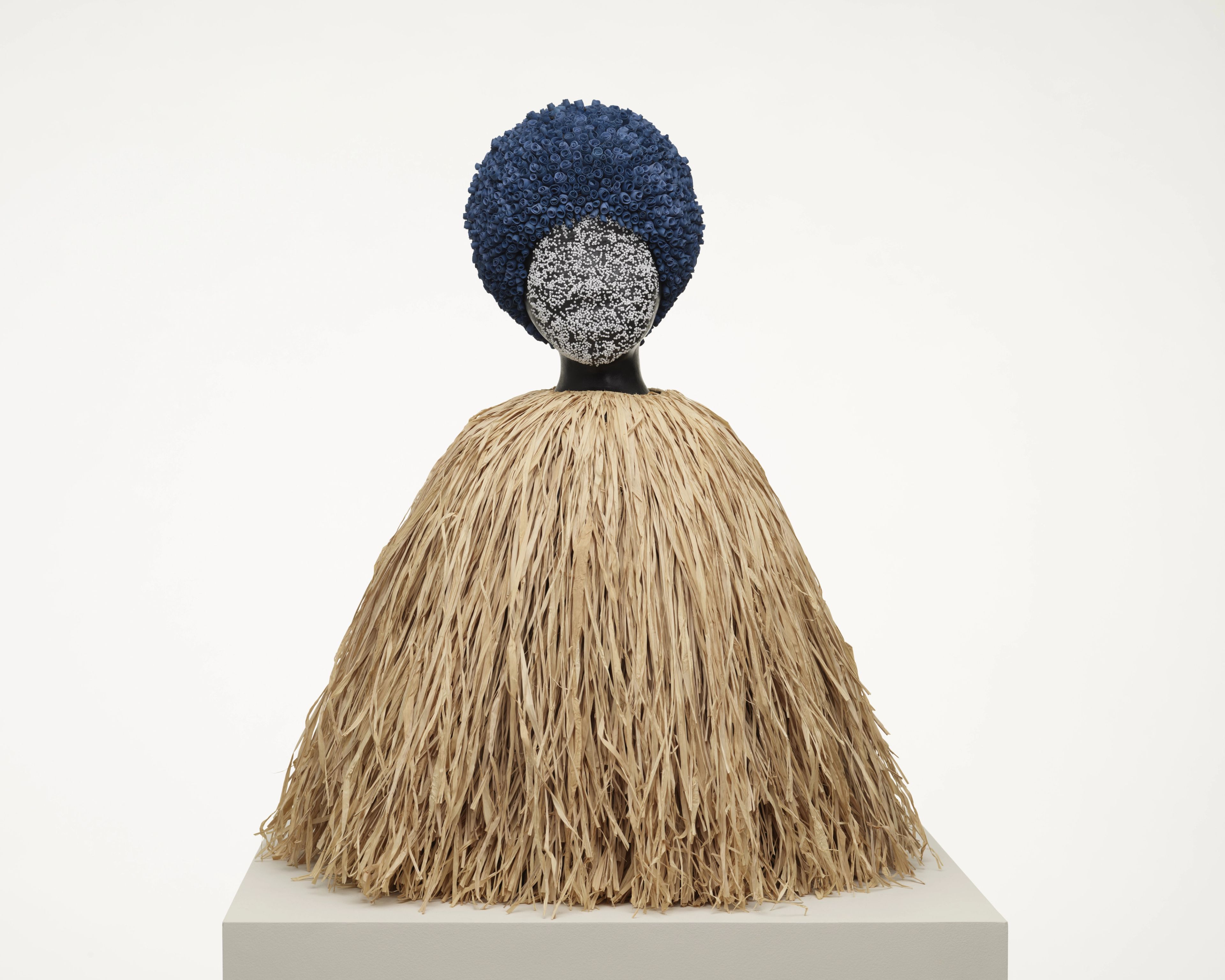 Figurative sculpture with a textured blue afro, a speckled face, and a conical skirt of natural straw.