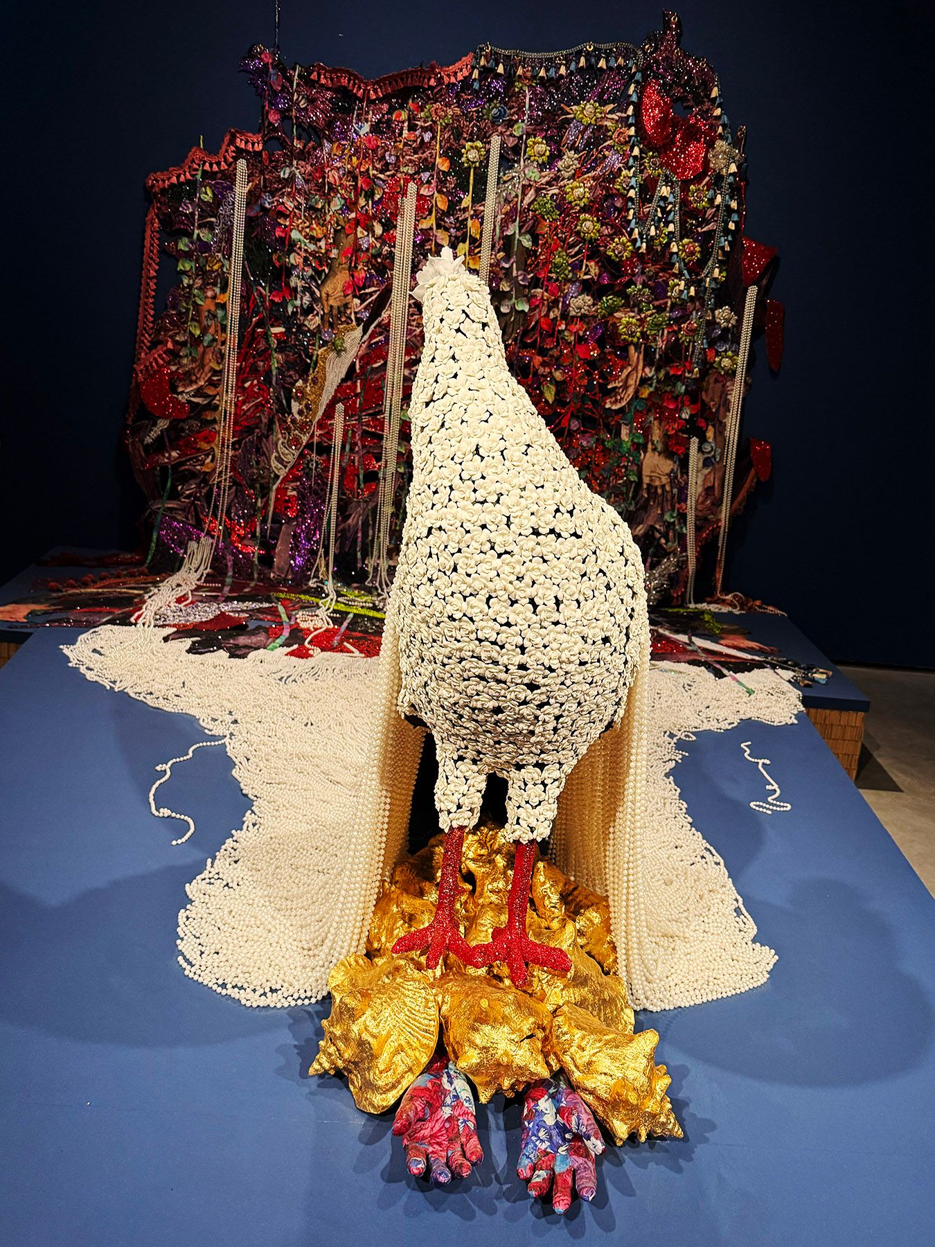 A white beaded bird sculpture with red feet stands on golden shells, draped with pearls, in front of a vibrant, ornate art installation.