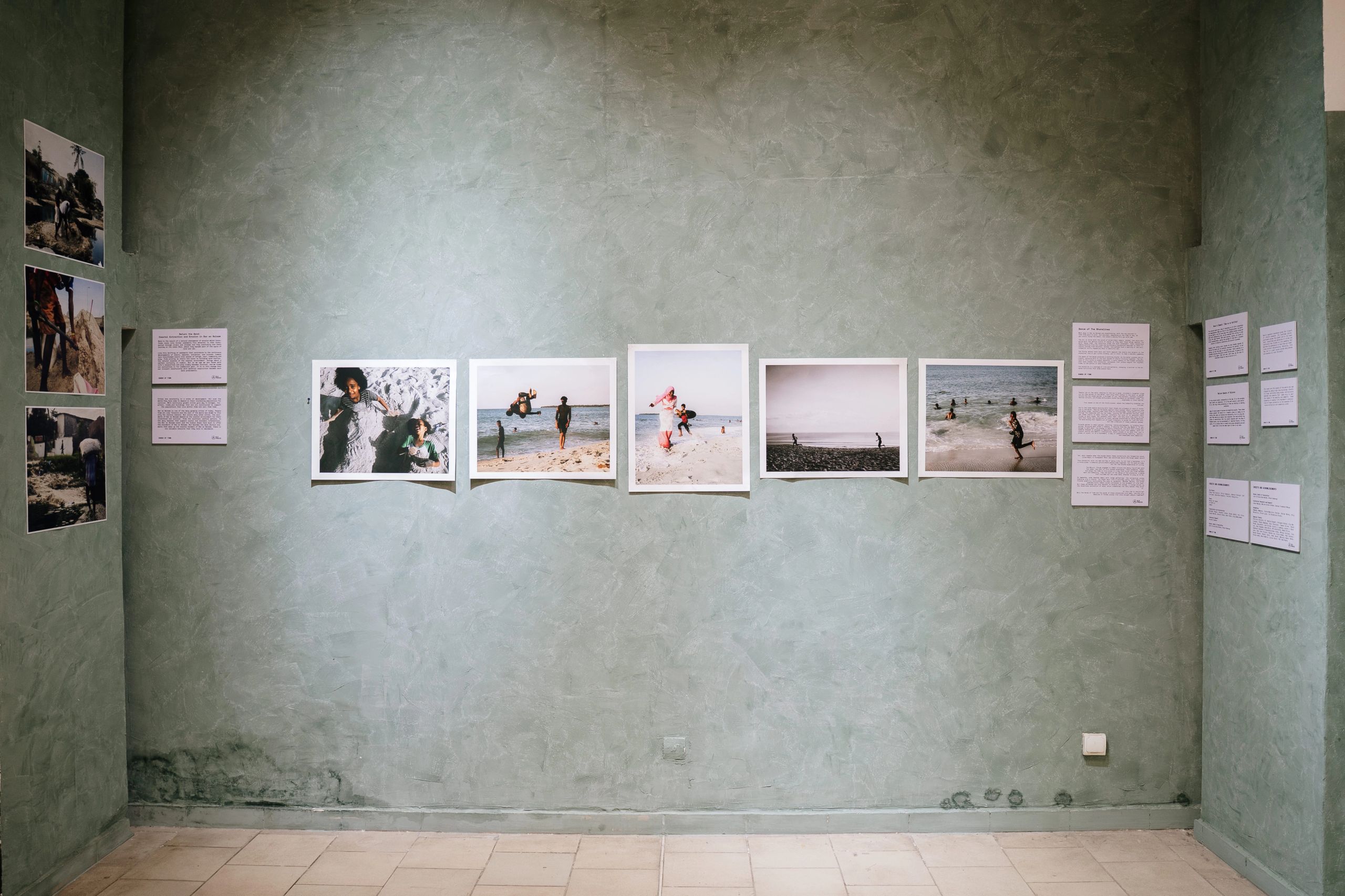 An art exhibition on a textured green wall featuring framed photographs of people on beaches and several text panels.