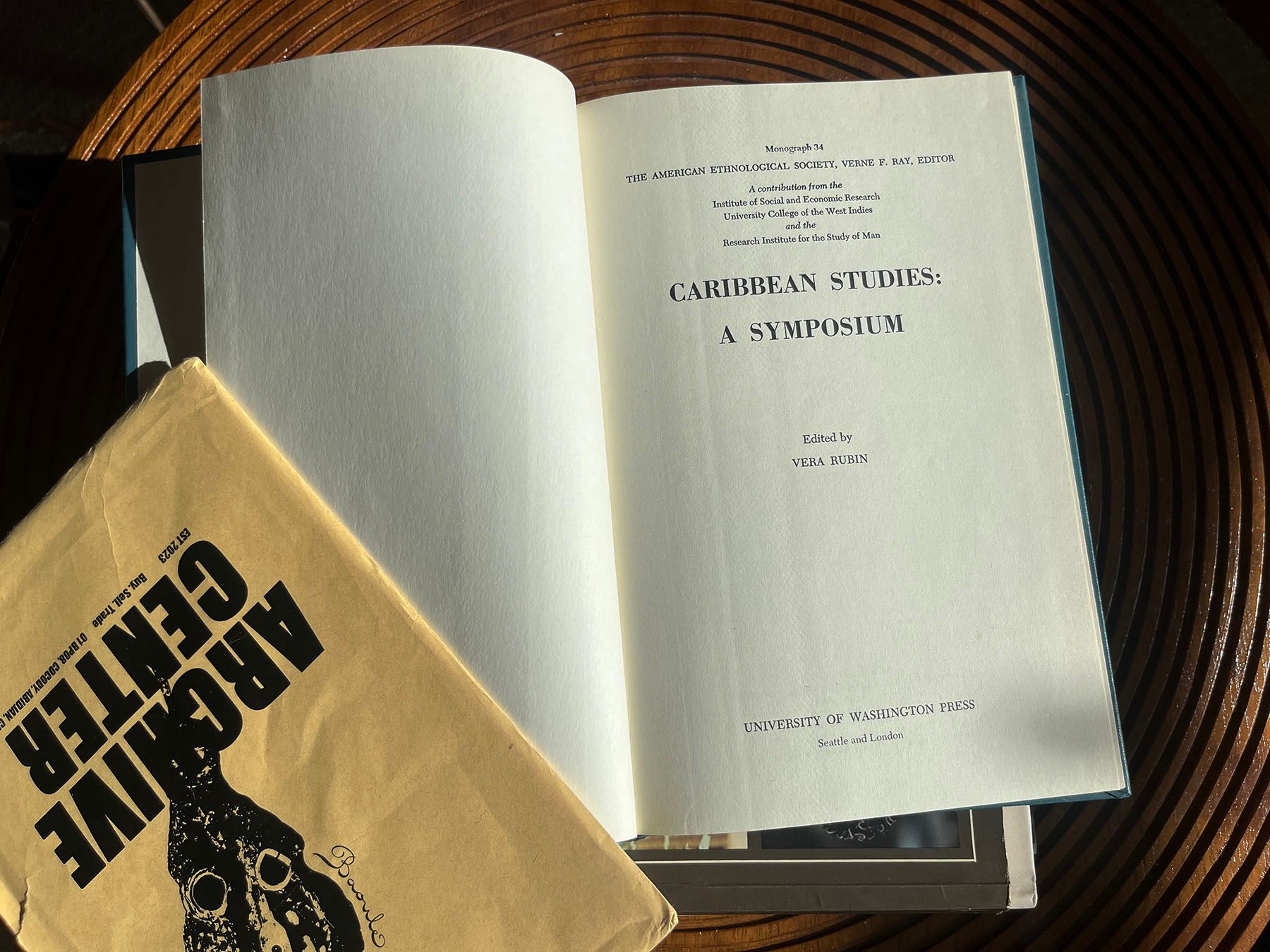 An open book titled "Caribbean Studies: A Symposium" edited by Vera Rubin, next to an "Archive Center" folder, on a wooden surface.