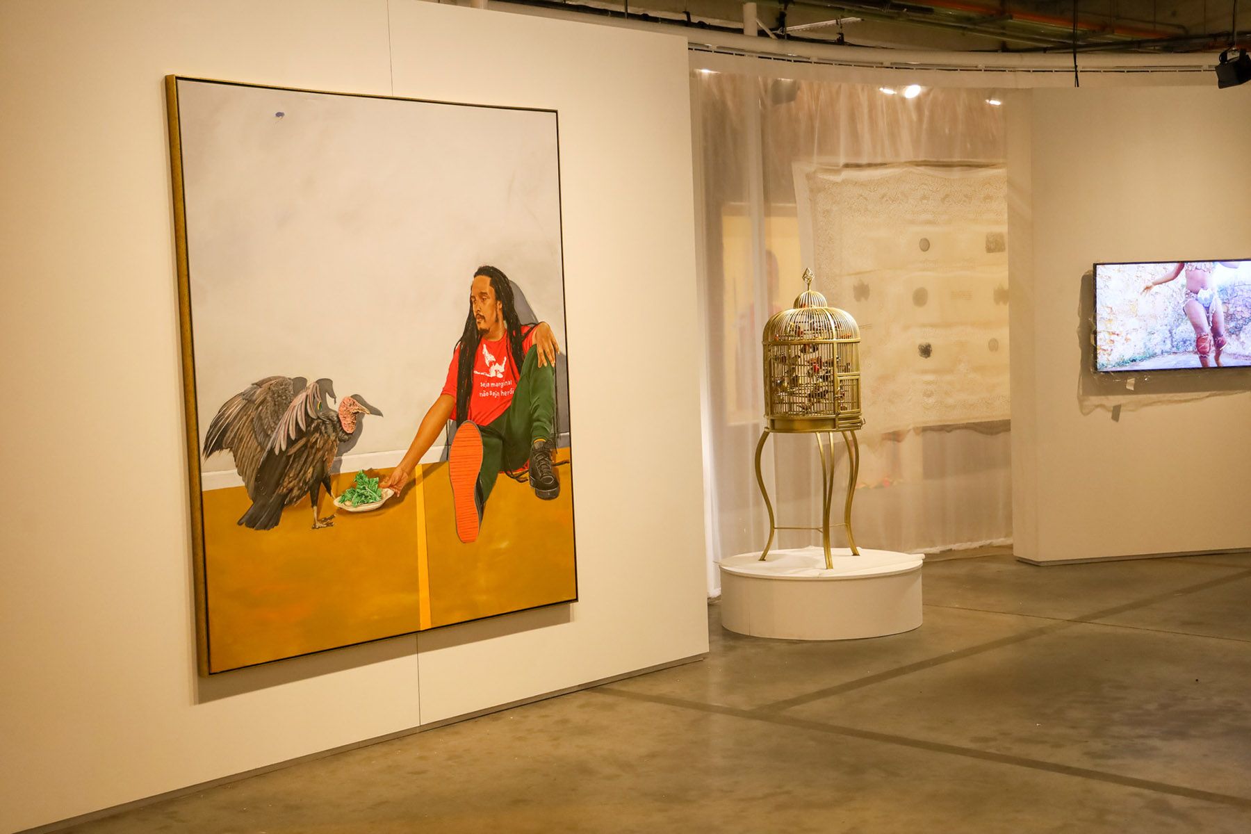 Art gallery with a painting of a person feeding a vulture, a birdcage sculpture, and a video display.
