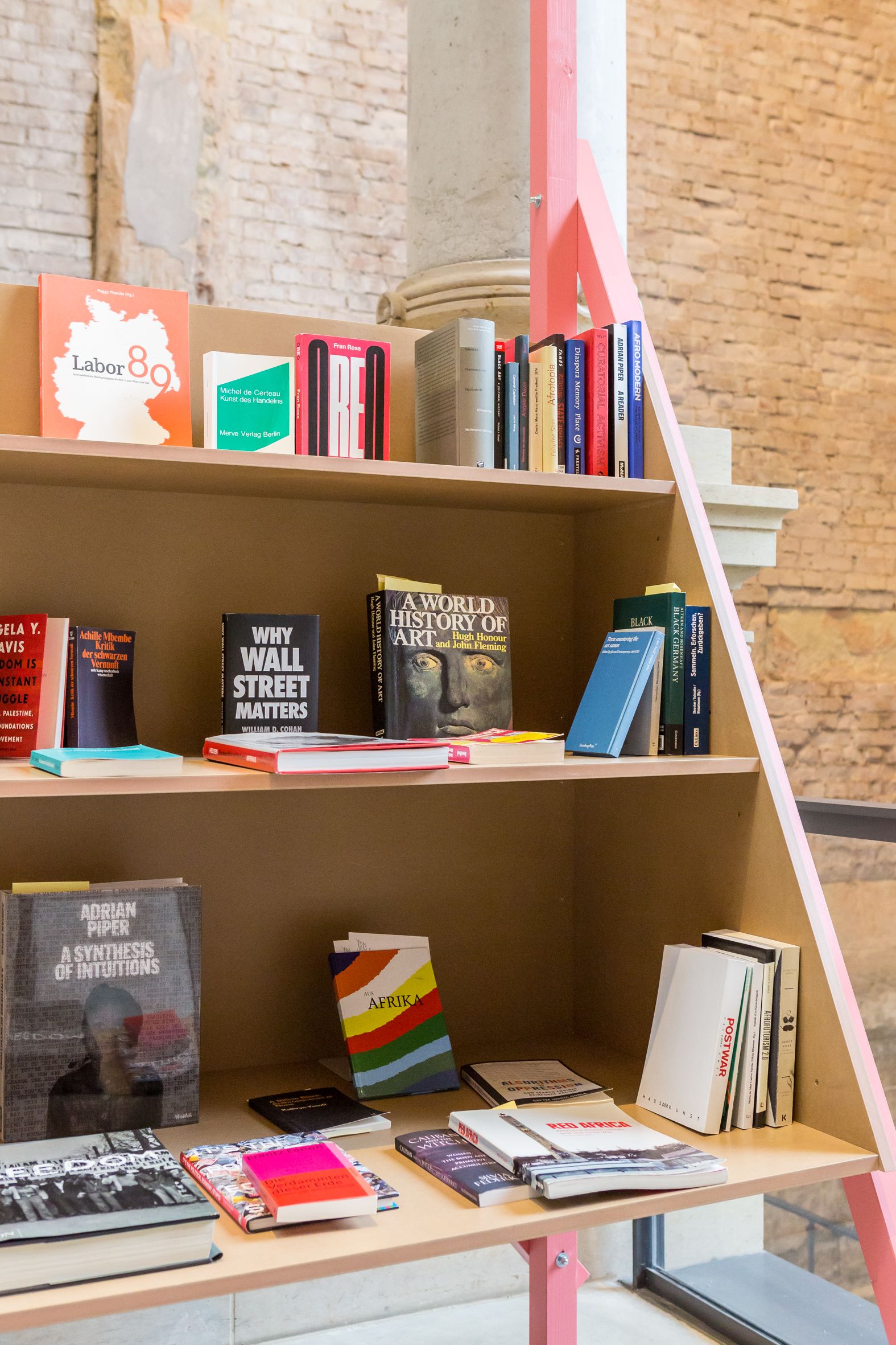 A light wooden bookshelf filled with books, with a bright pink triangular support on the right, against a brick wall and a stone column.