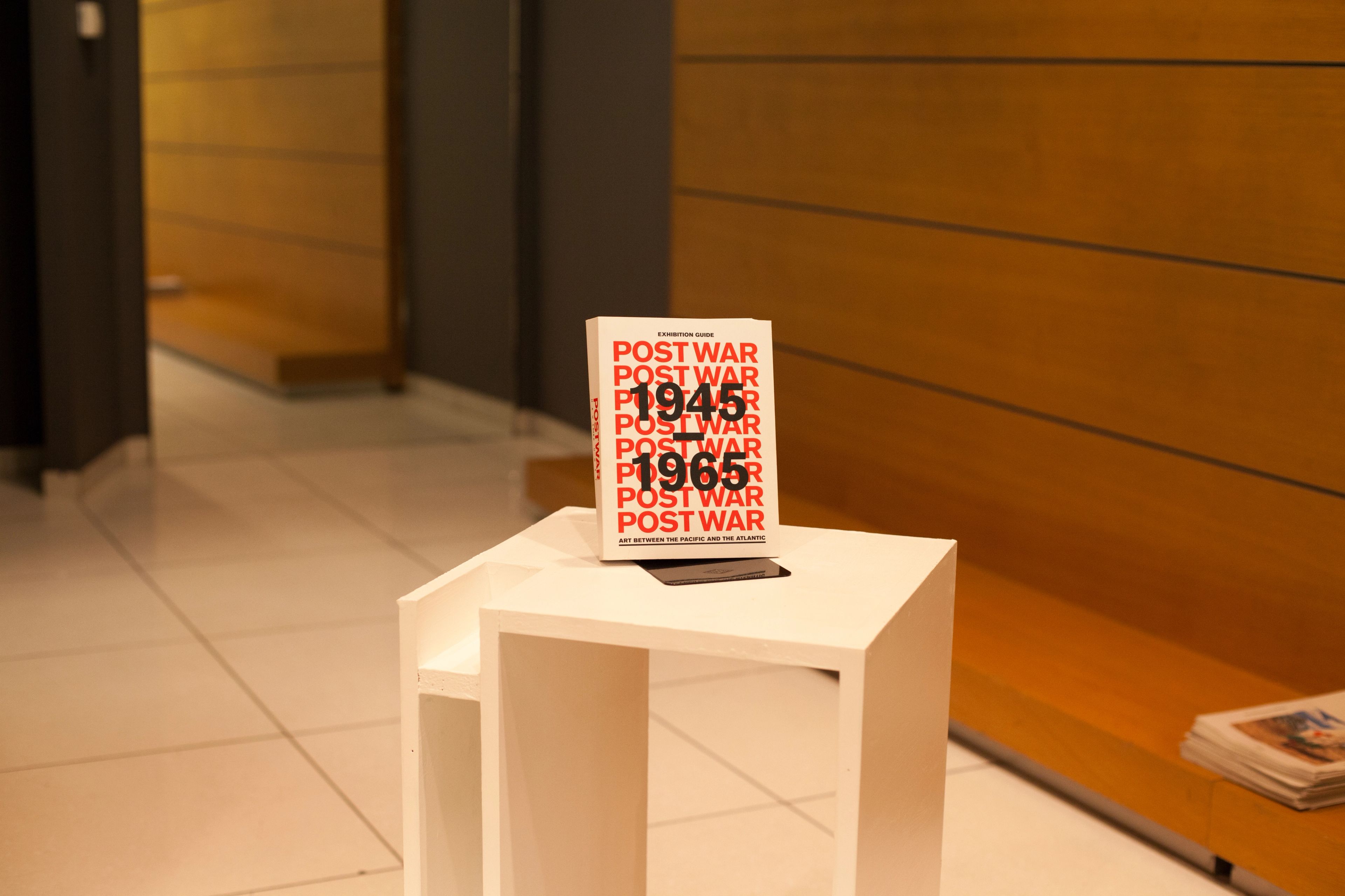 A white book with "POST WAR 1945 1965" in red text stands on a small white table in a modern interior.