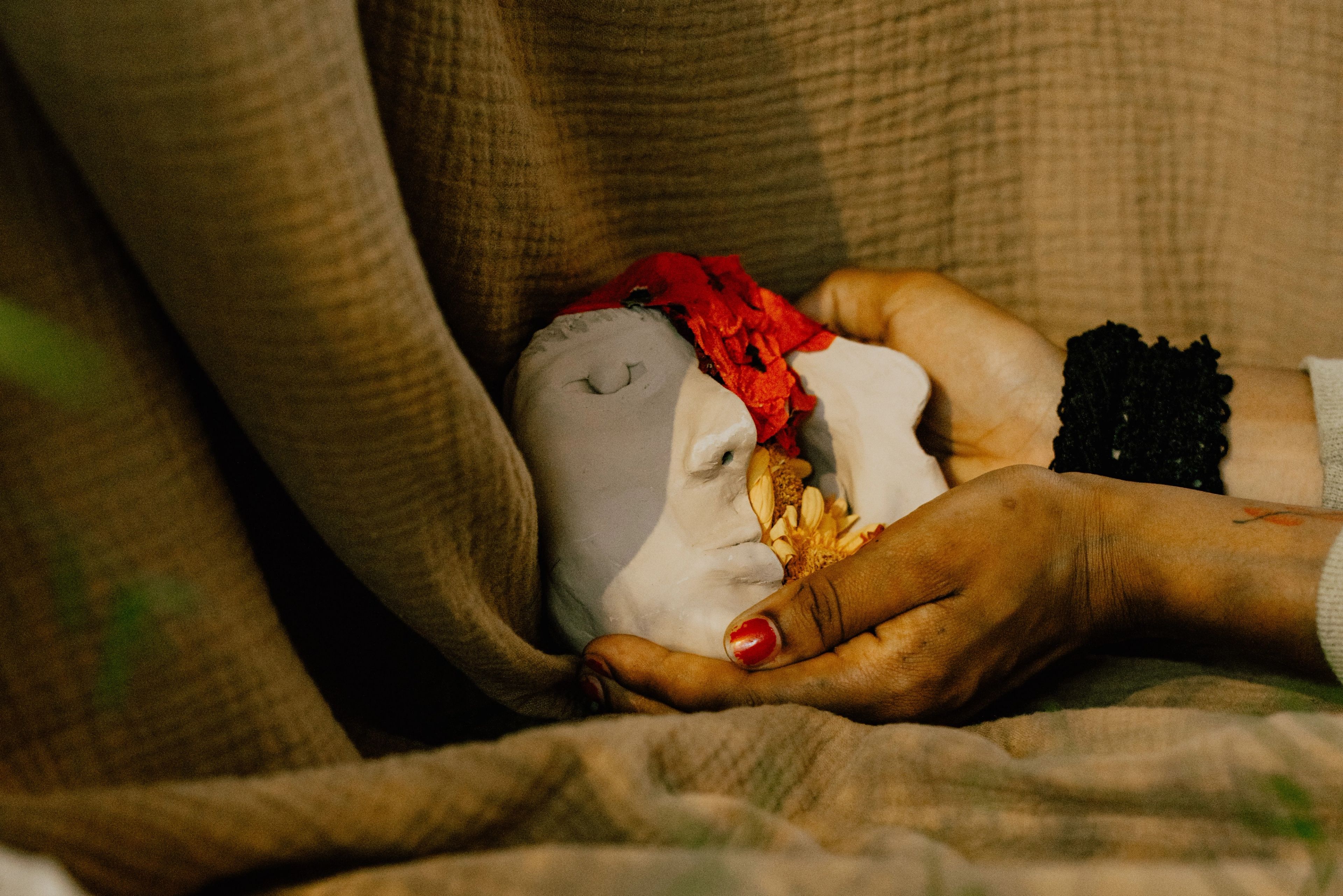 Hands holding a broken white face mask filled with red petals and a dried yellow flower.