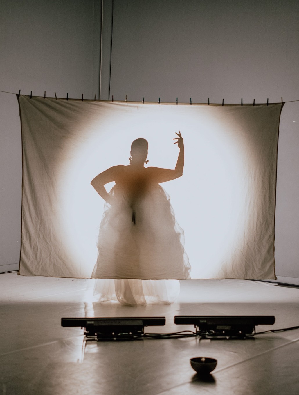 Backlit silhouette of a person in a voluminous dress behind a screen, with sensors and a bowl on the floor.