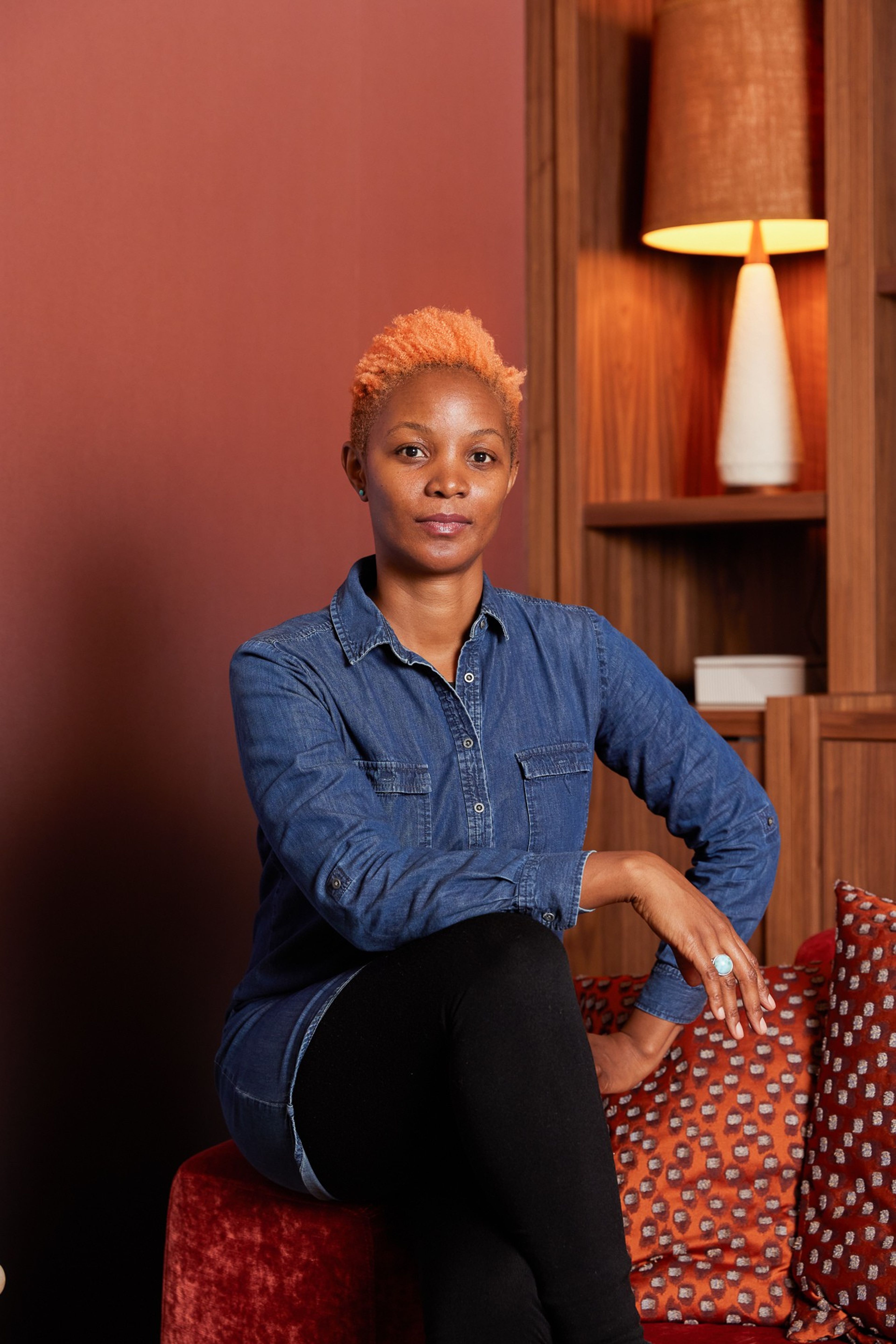 A woman with short orange hair, wearing a denim shirt, sits and looks at the camera.