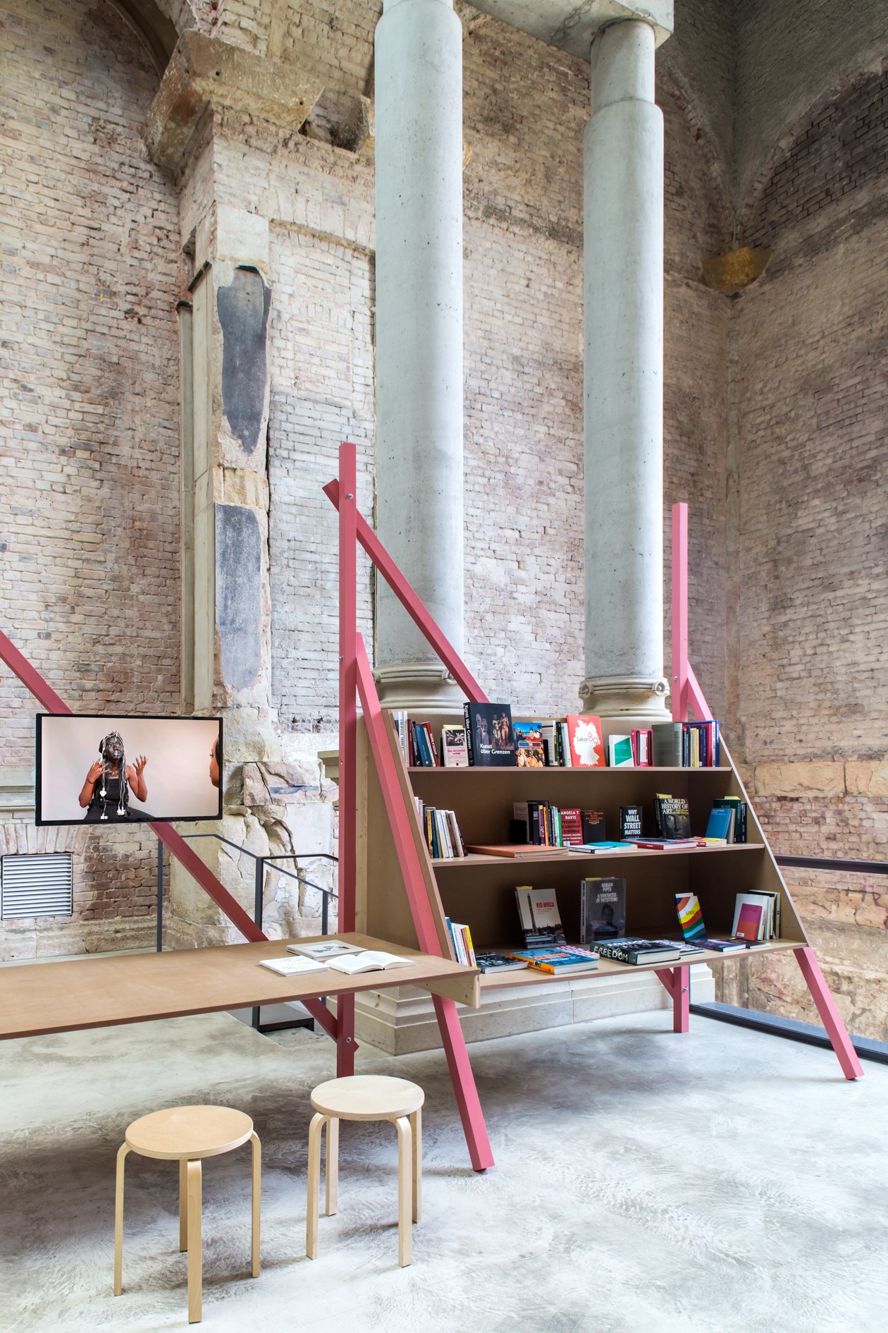 A large pink-framed bookshelf and desk with two stools stand in a brick-walled room with concrete pillars, next to a TV showing a person in a VR headset.
