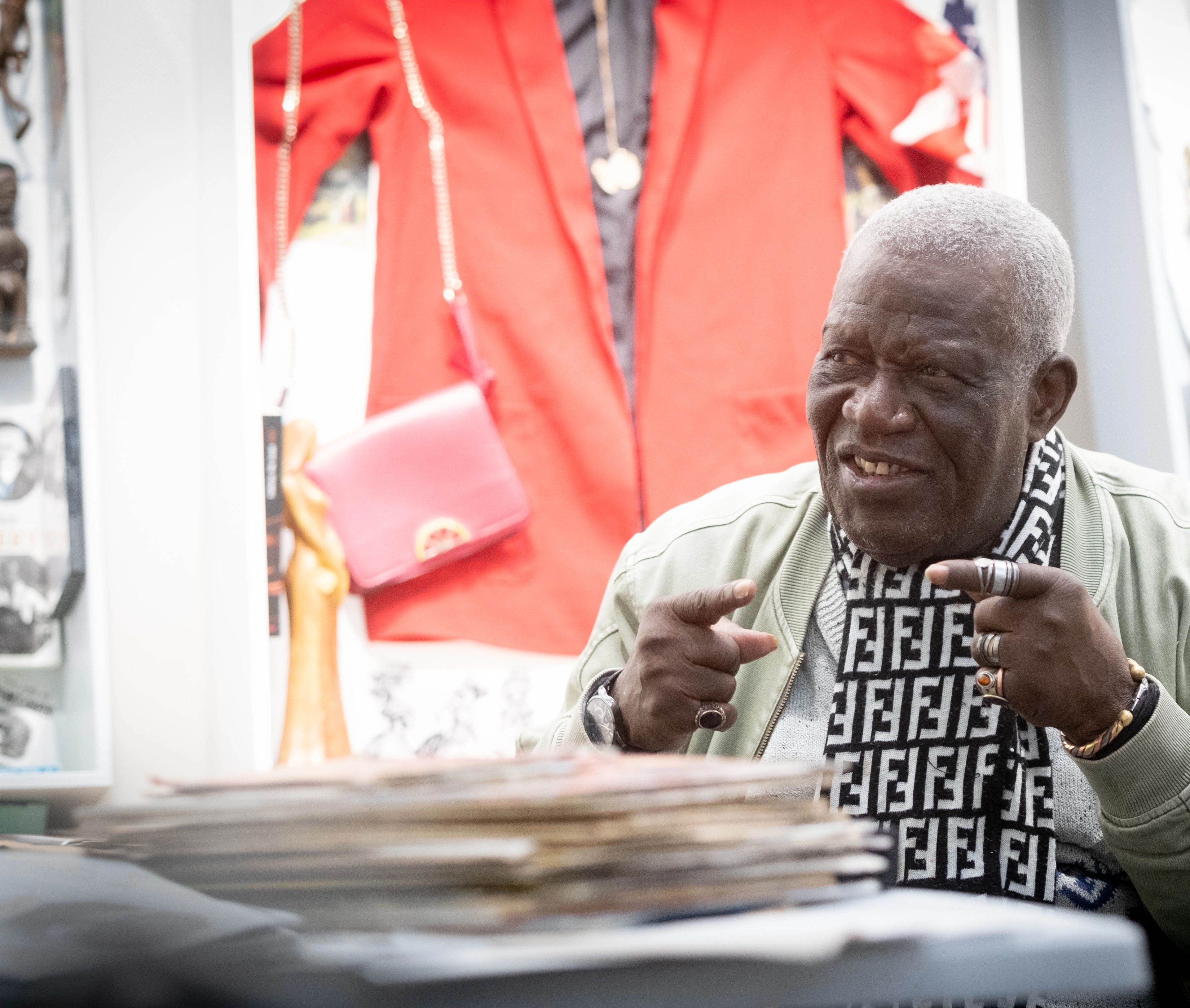 An older Black man wearing a Fendi scarf gestures with both hands, with a red jacket and pink purse in the blurred background.