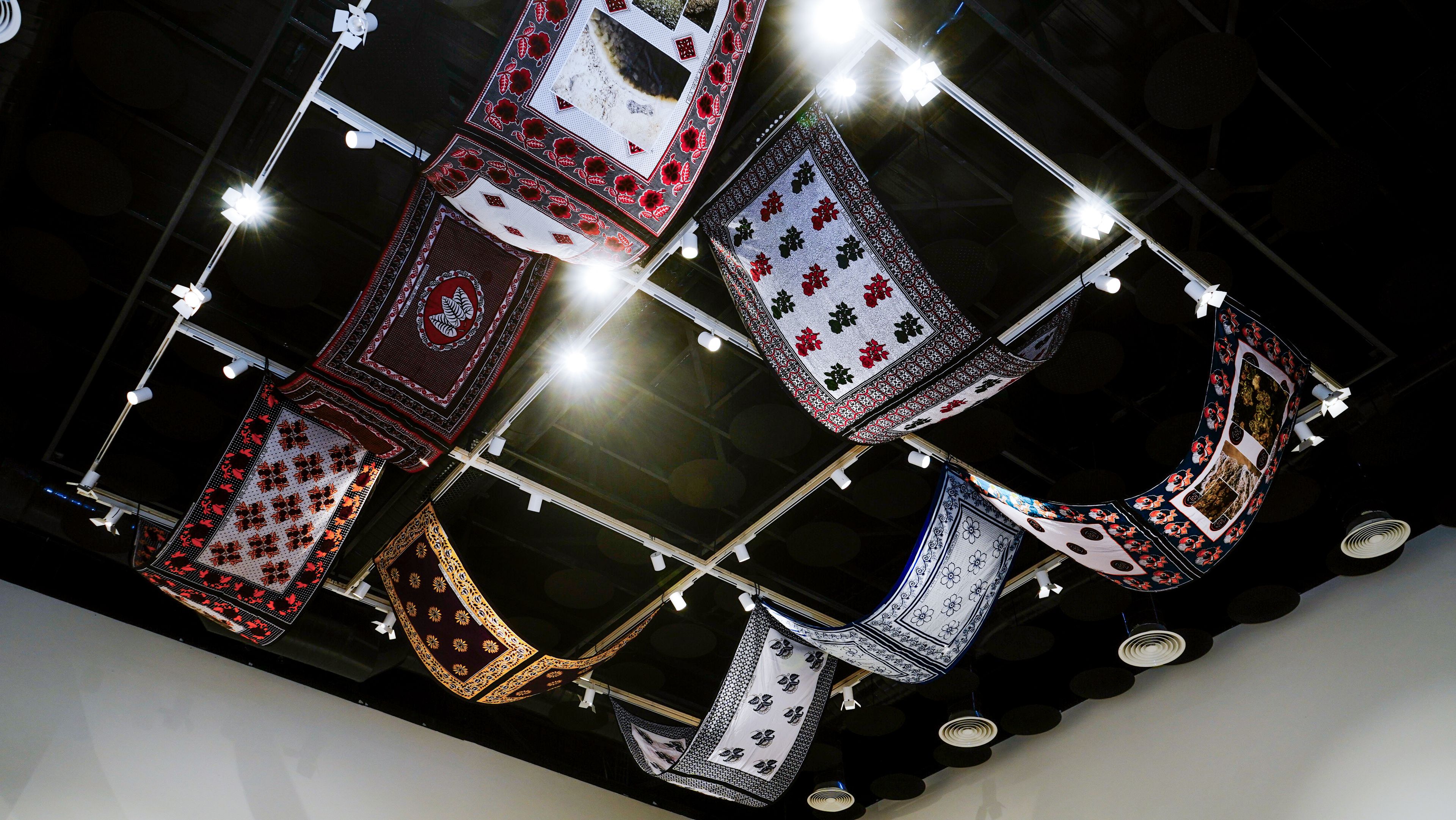 Patterned textiles hang in undulating waves from a dark ceiling, illuminated by spotlights.
