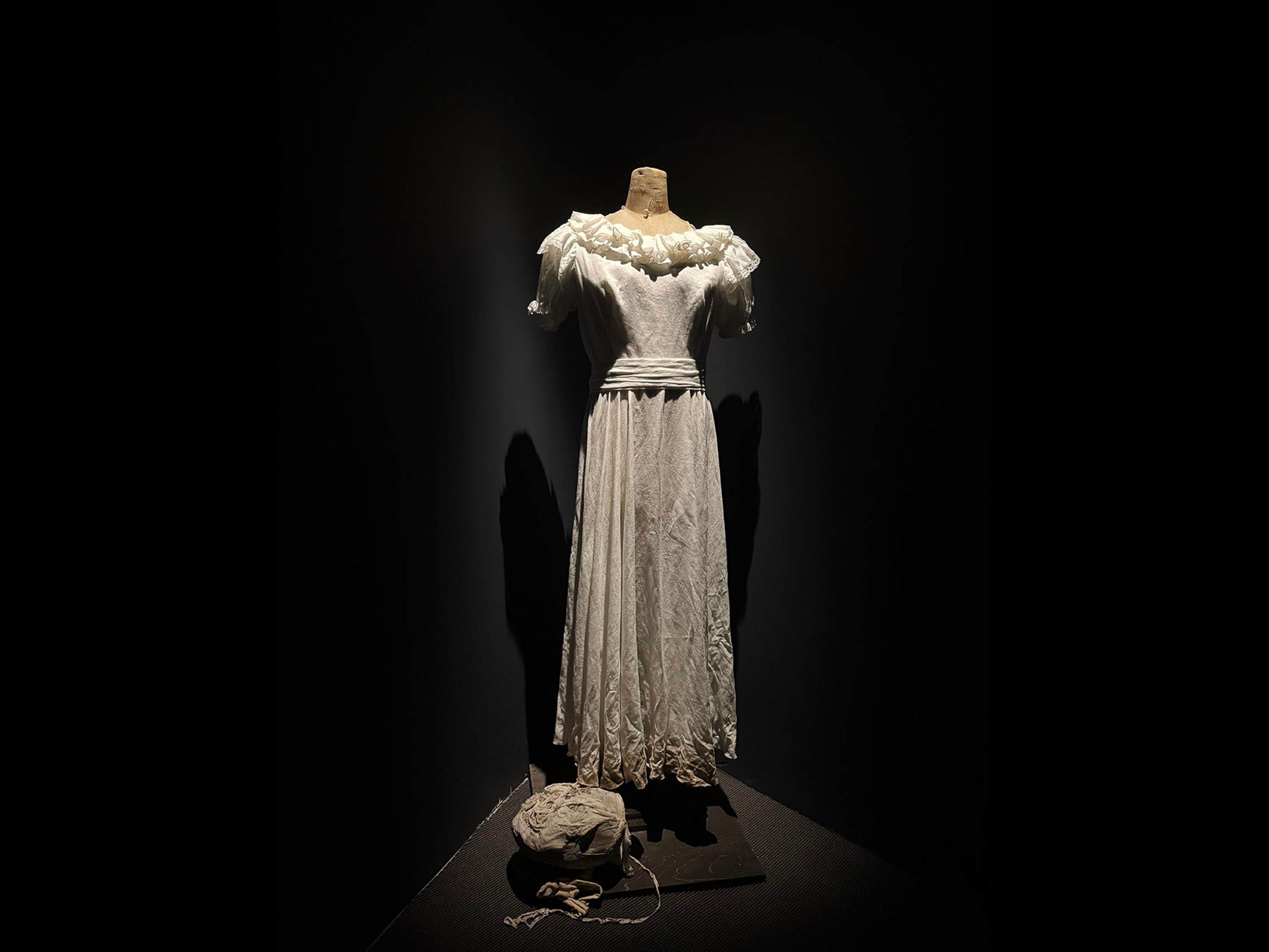 A white, ruffled vintage dress on a headless mannequin, brightly lit against a dark background. A crumpled white object lies on the floor.