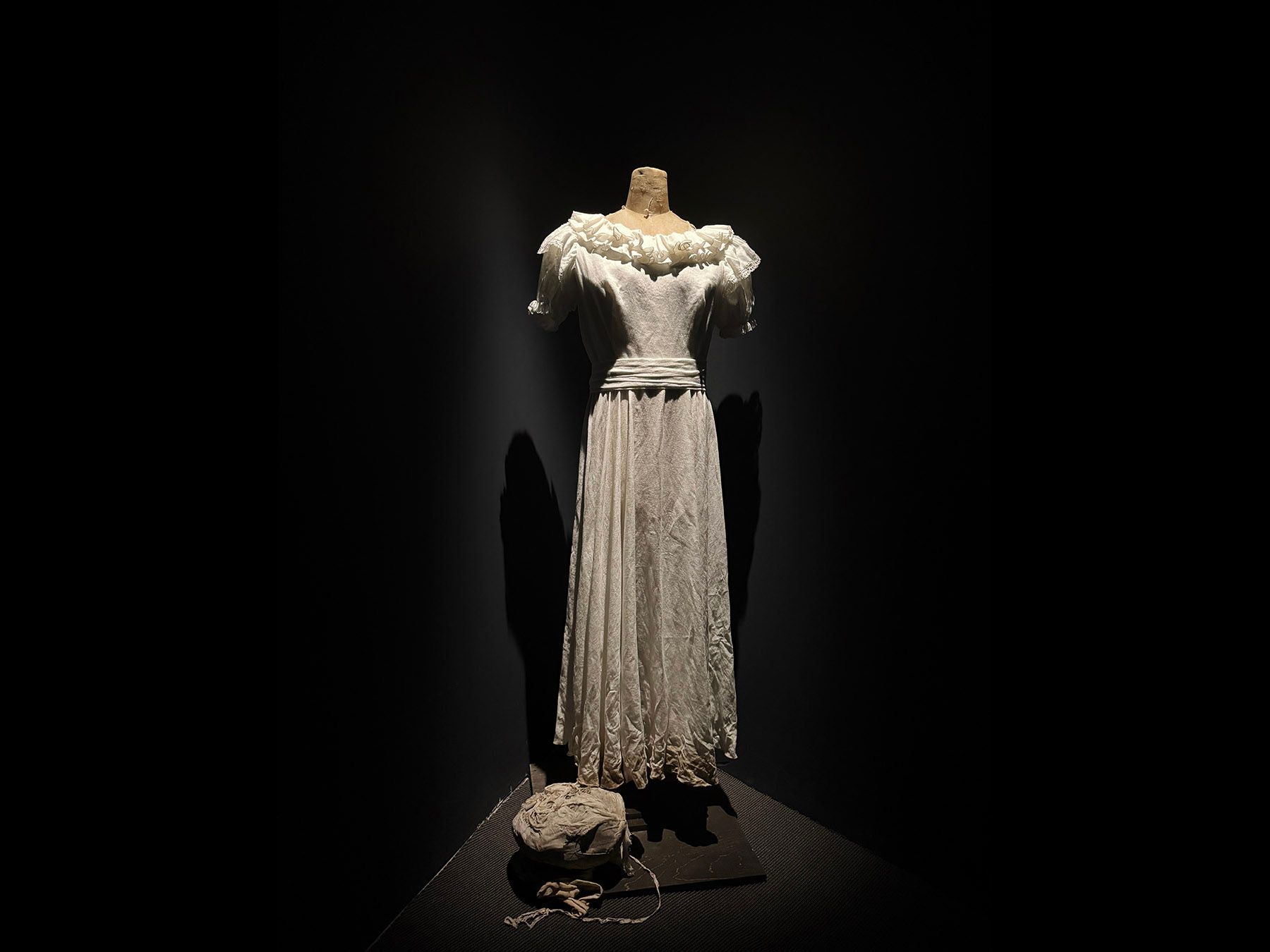 A white, ruffled vintage dress on a headless mannequin, brightly lit against a dark background. A crumpled white object lies on the floor.