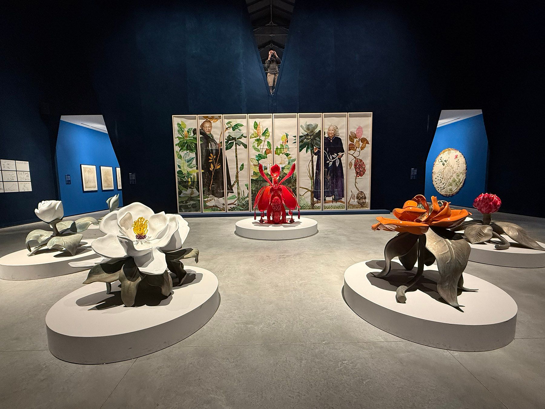 Art exhibition featuring large colorful flower sculptures on white pedestals, with multi-panel botanical illustrations and figures in the background.