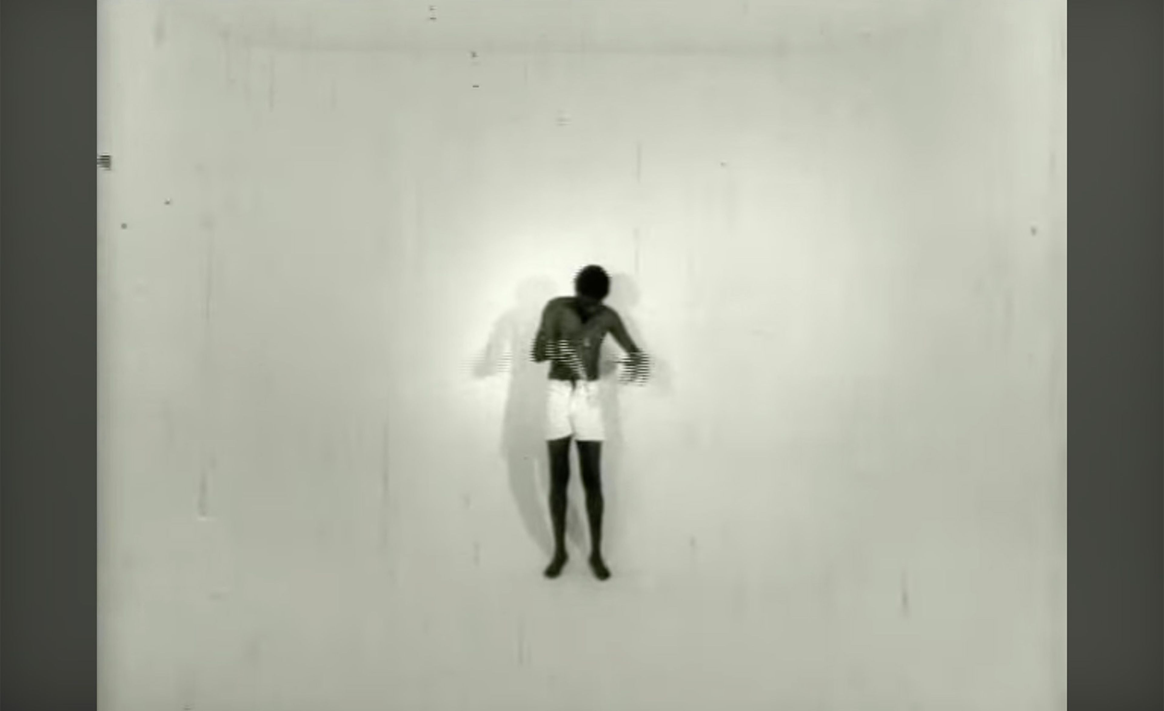 Black and white still of a shirtless, dark-skinned person in white shorts, hands together, standing centrally in a stark white room with a shadow.