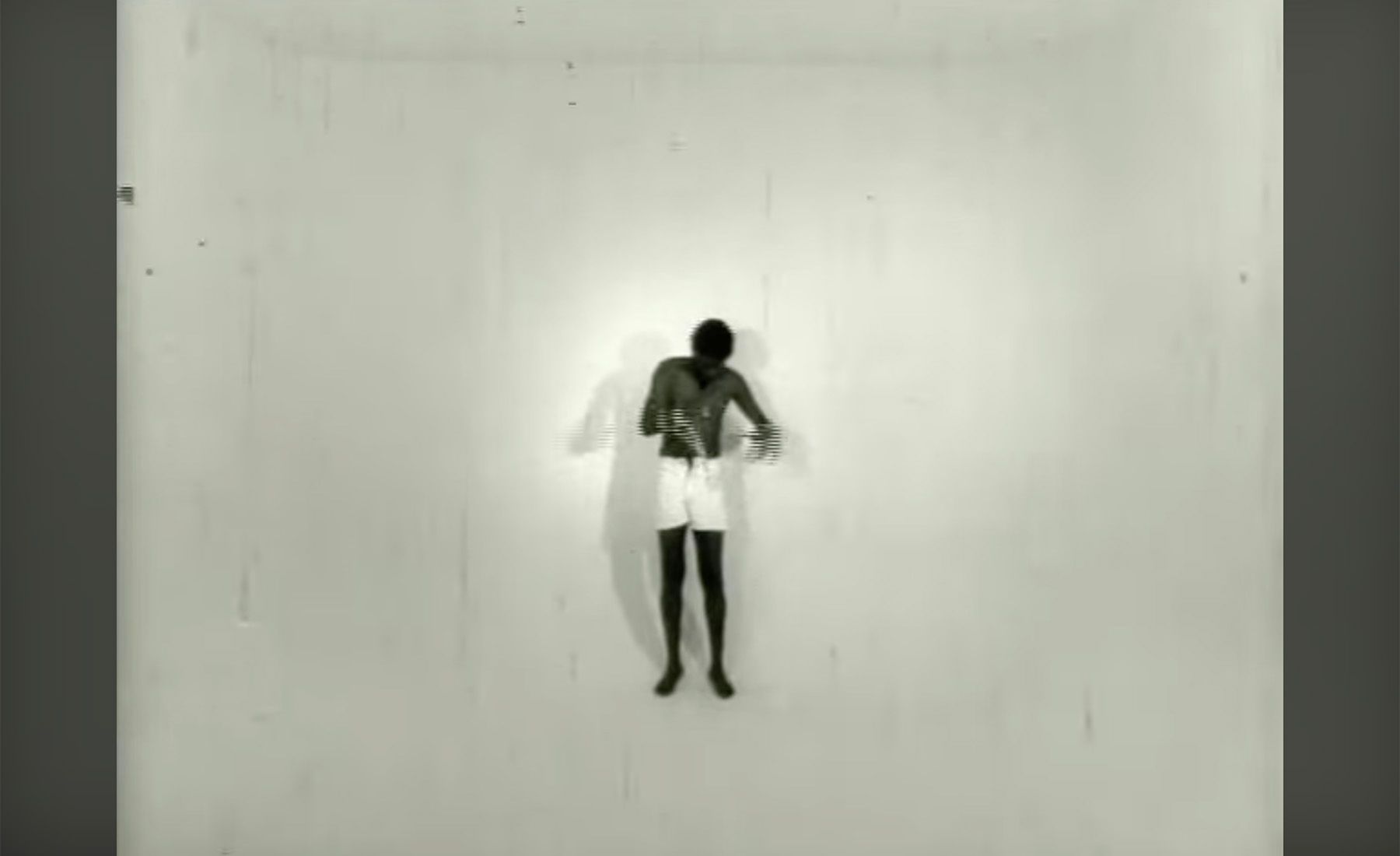 Black and white still of a shirtless, dark-skinned person in white shorts, hands together, standing centrally in a stark white room with a shadow.