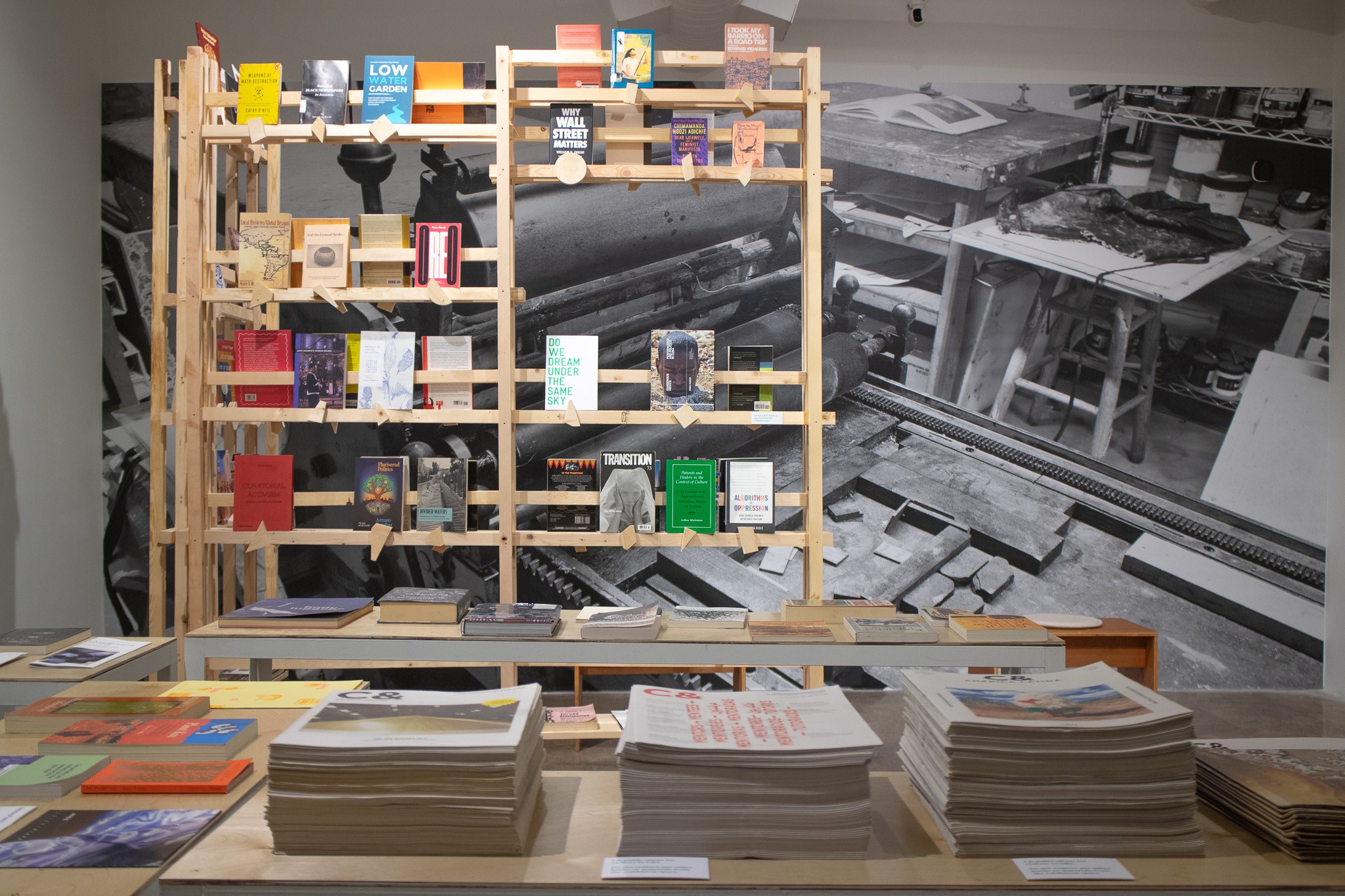 A gallery display with books on a wooden grid shelf, a black and white photo mural of a workshop, and stacks of publications on foreground tables.