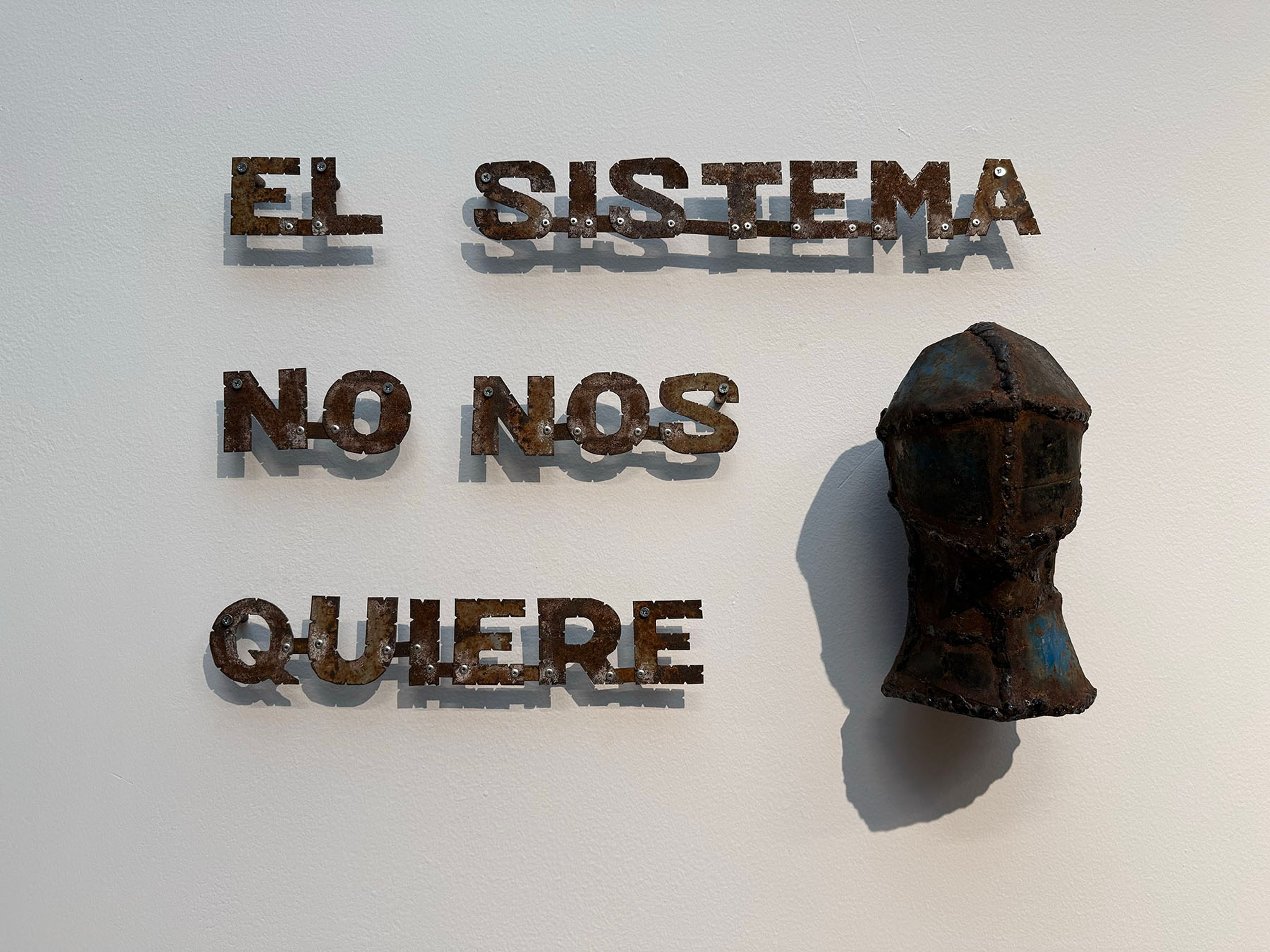 Art installation with rusty metal letters spelling "EL SISTEMA NO NOS QUIERE" next to a dark, rusty, abstract head sculpture with blue accents on a white wall.
