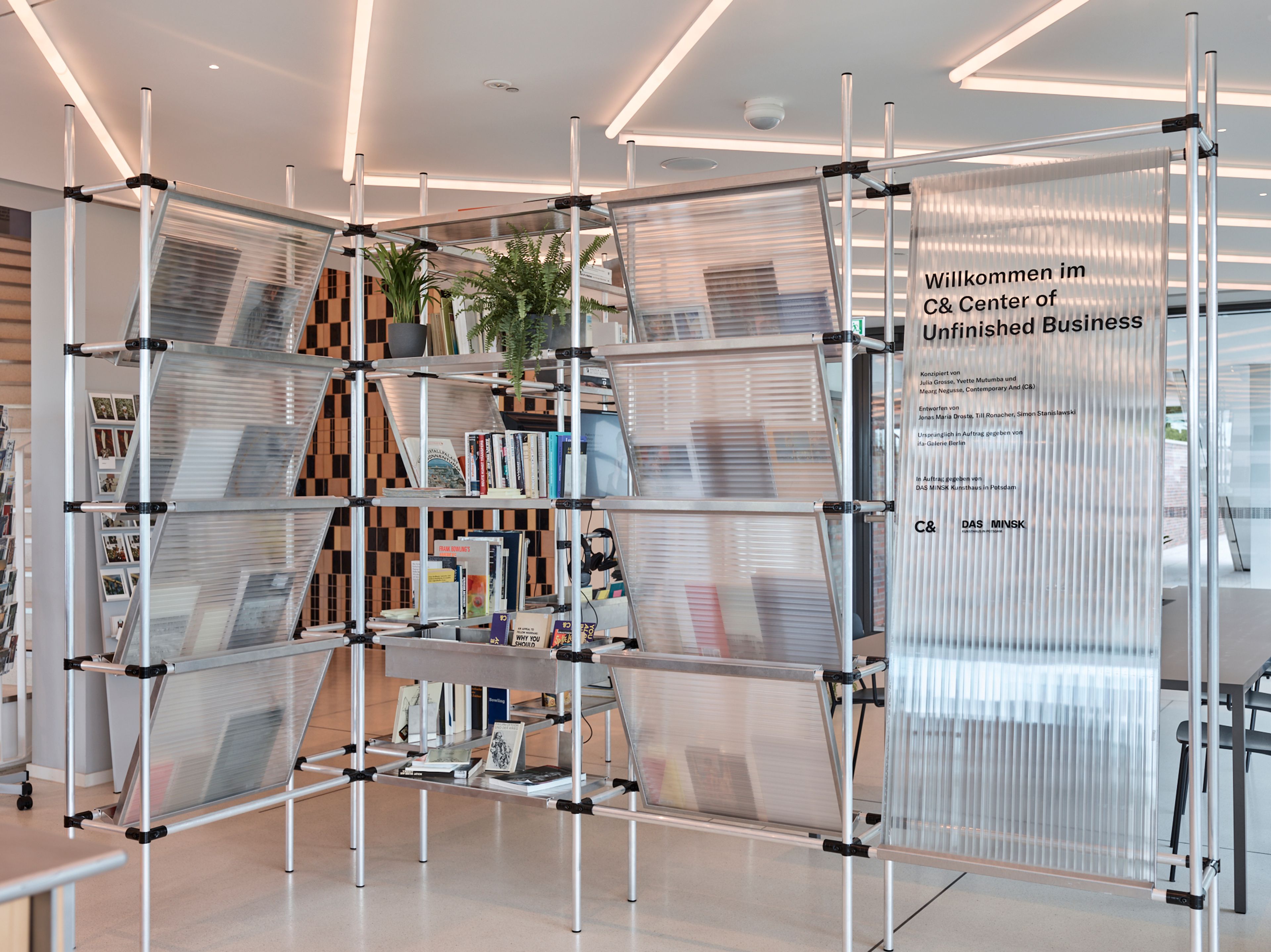 A modern modular shelving unit made of aluminum pipes and translucent panels, holding books, plants, and a sign that reads "Willkommen im C& Center of Unfinished Business."