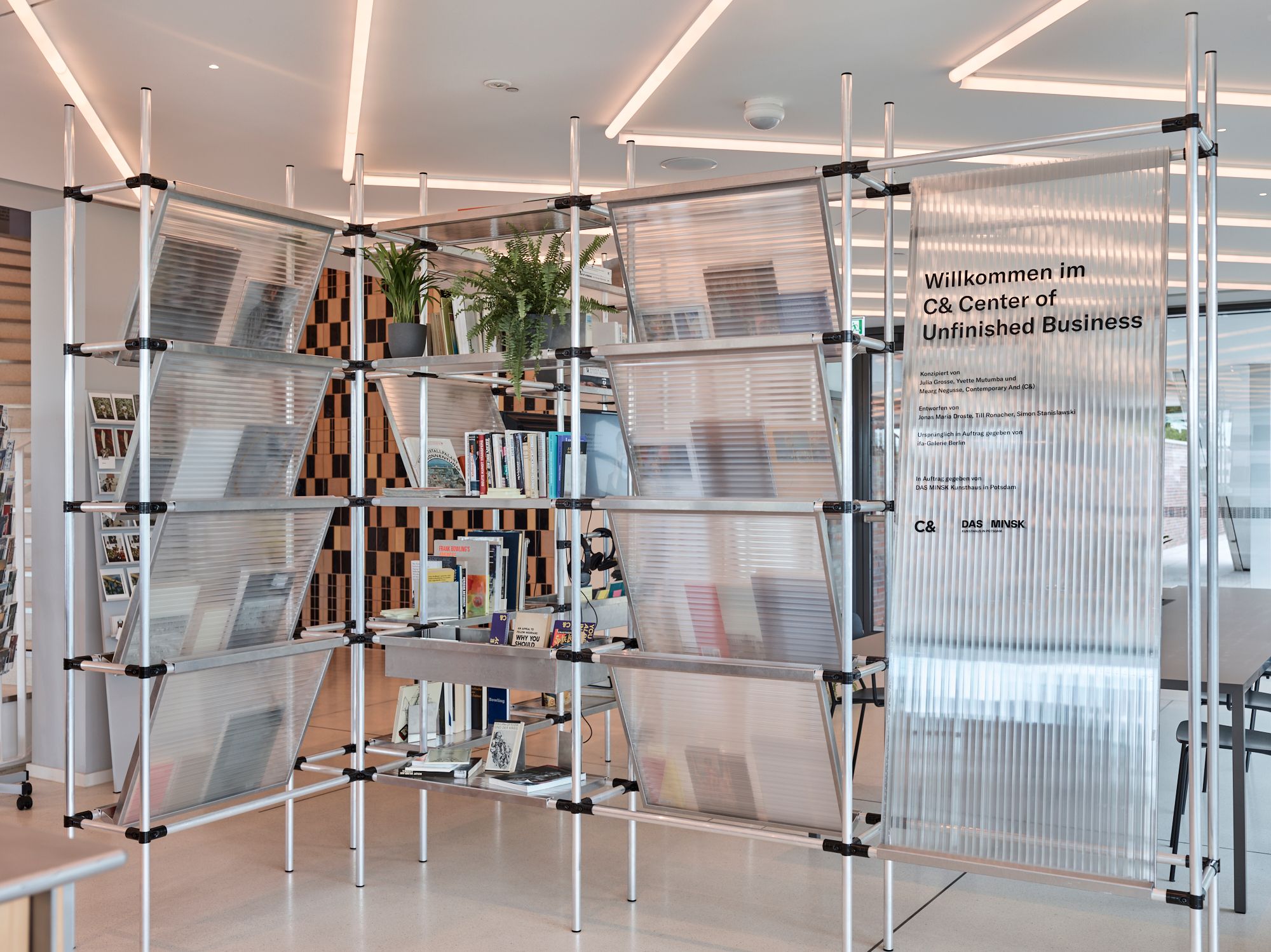 A modern modular shelving unit made of aluminum pipes and translucent panels, holding books, plants, and a sign that reads "Willkommen im C& Center of Unfinished Business."