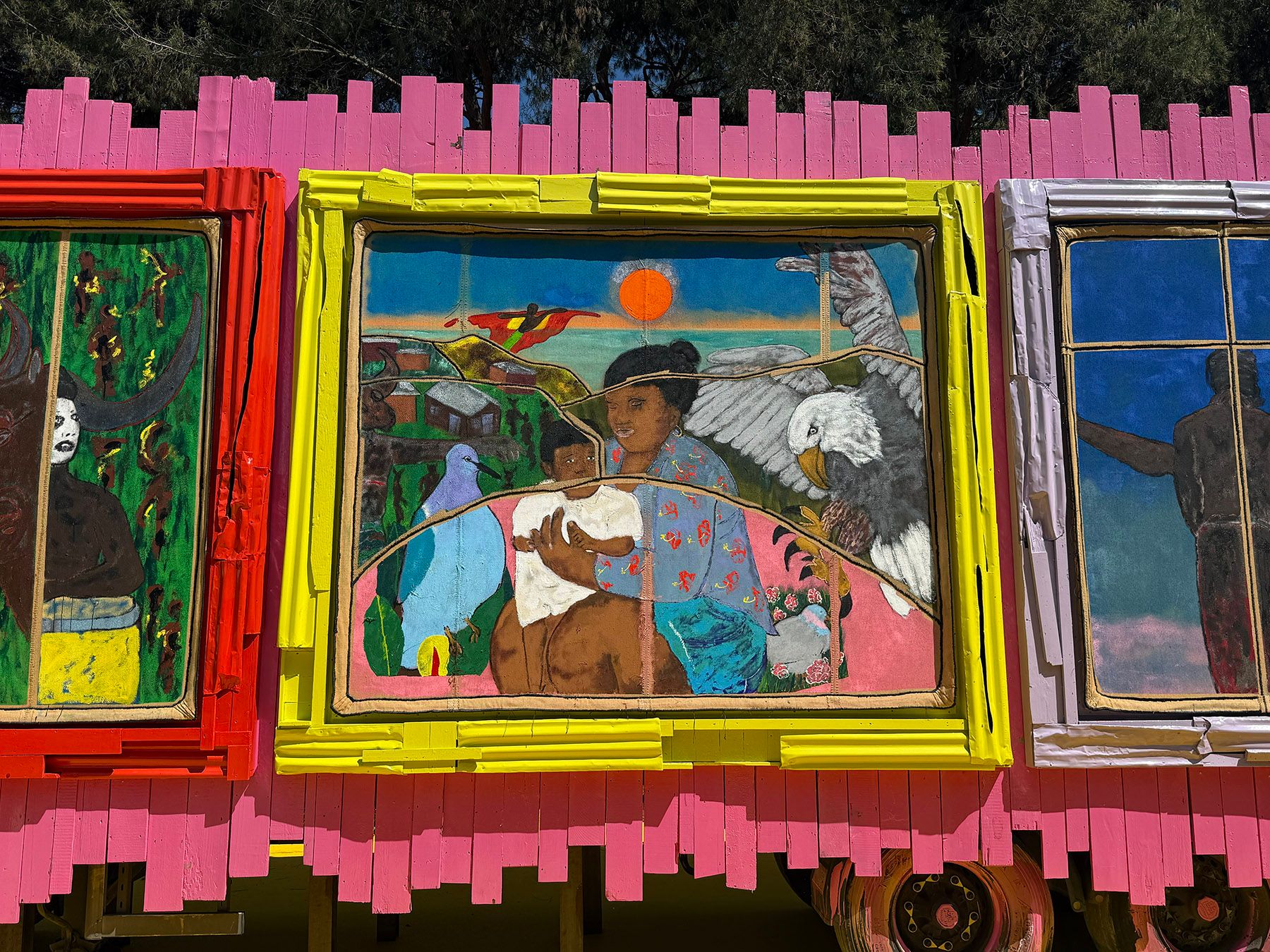 Three vibrant folk art paintings, the central one depicting a woman, child, blue bird, and eagle in a yellow frame, set against a pink wooden wall.
