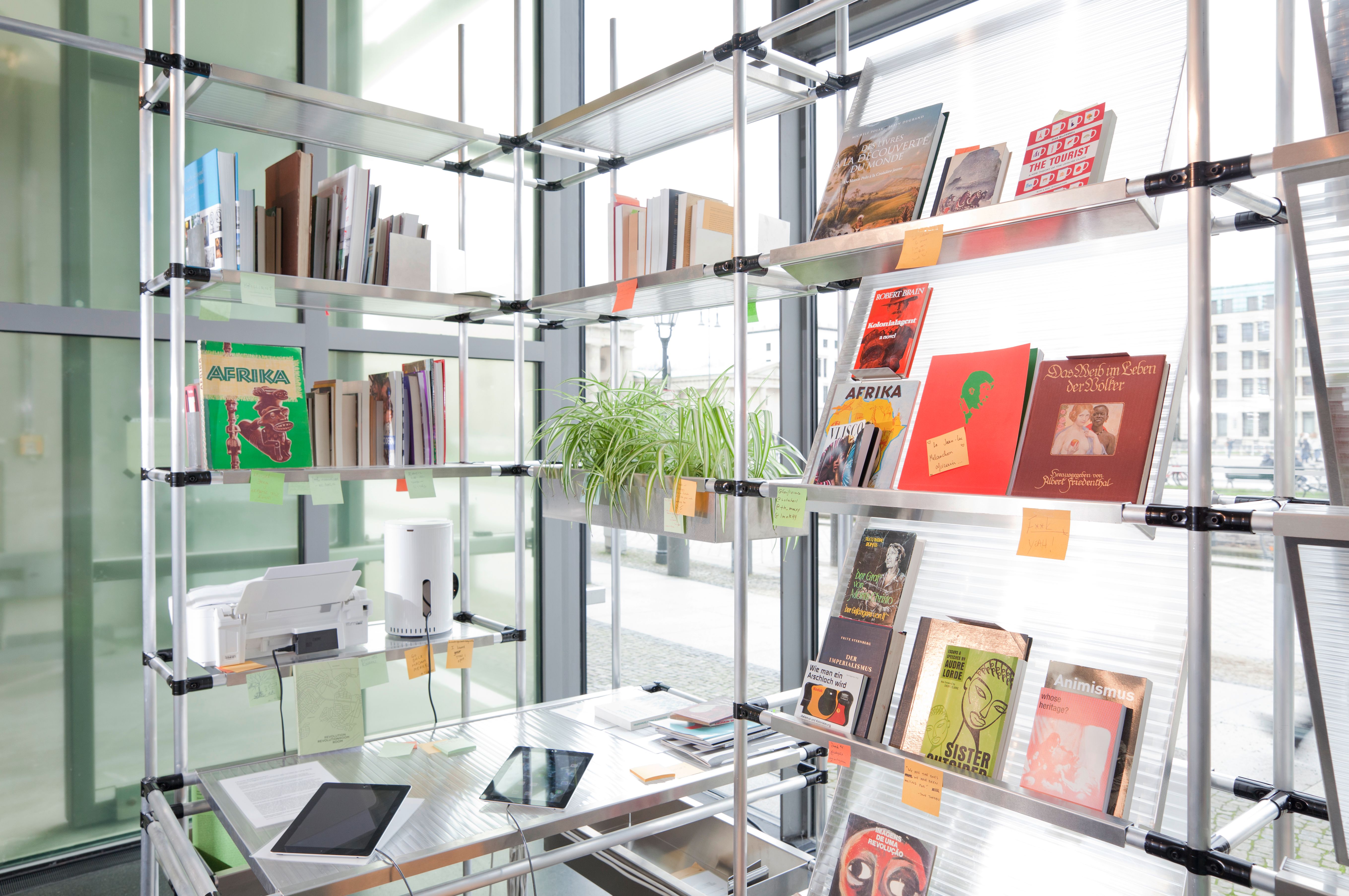 A modular shelving unit displaying books, magazines, a plant, a printer, and tablets, in front of large windows.