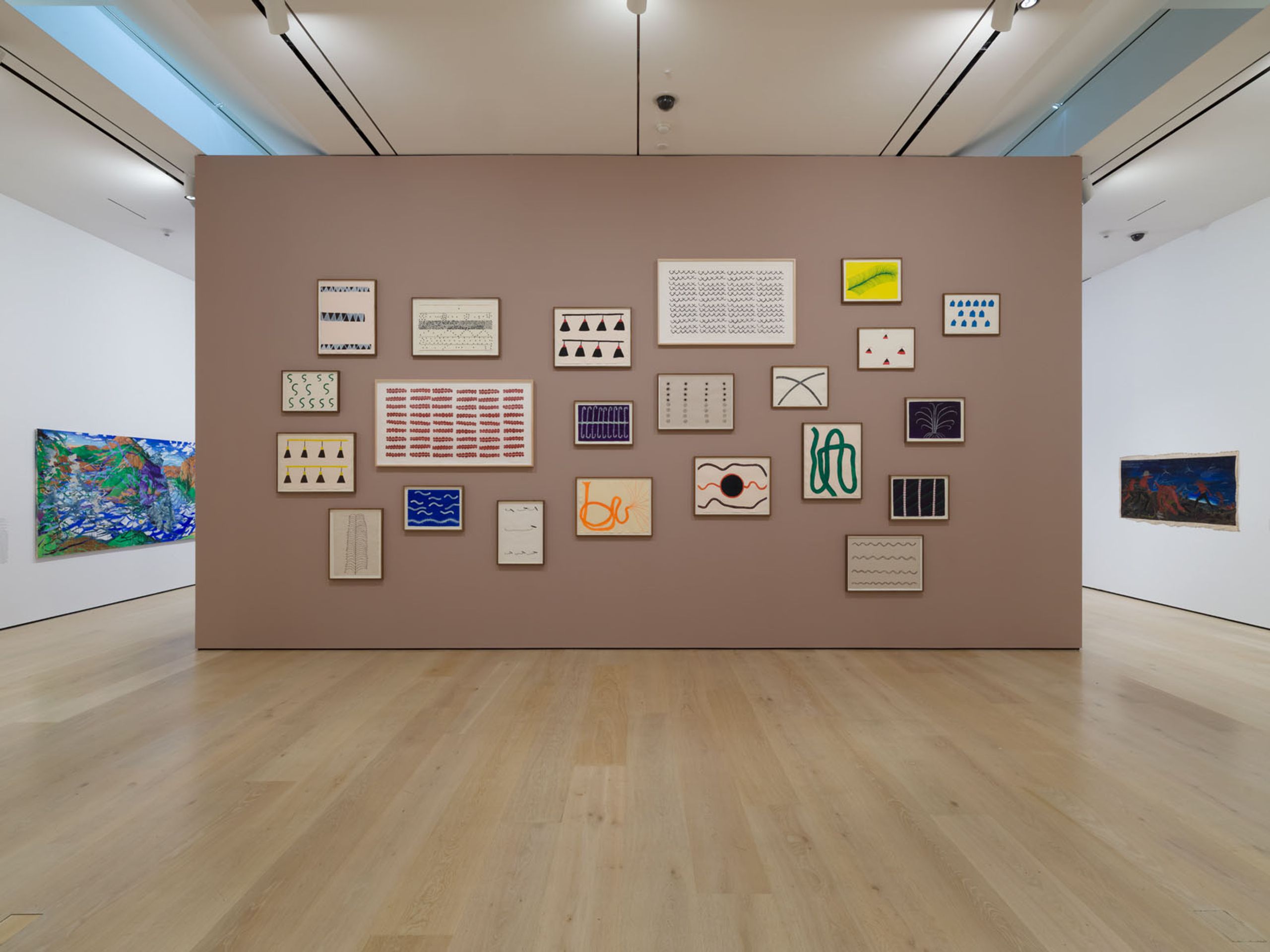An art gallery with a large taupe wall displaying numerous framed abstract artworks, flanked by two large colorful paintings on white walls.