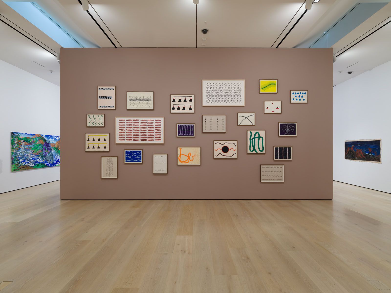 An art gallery with a large taupe wall displaying numerous framed abstract artworks, flanked by two large colorful paintings on white walls.