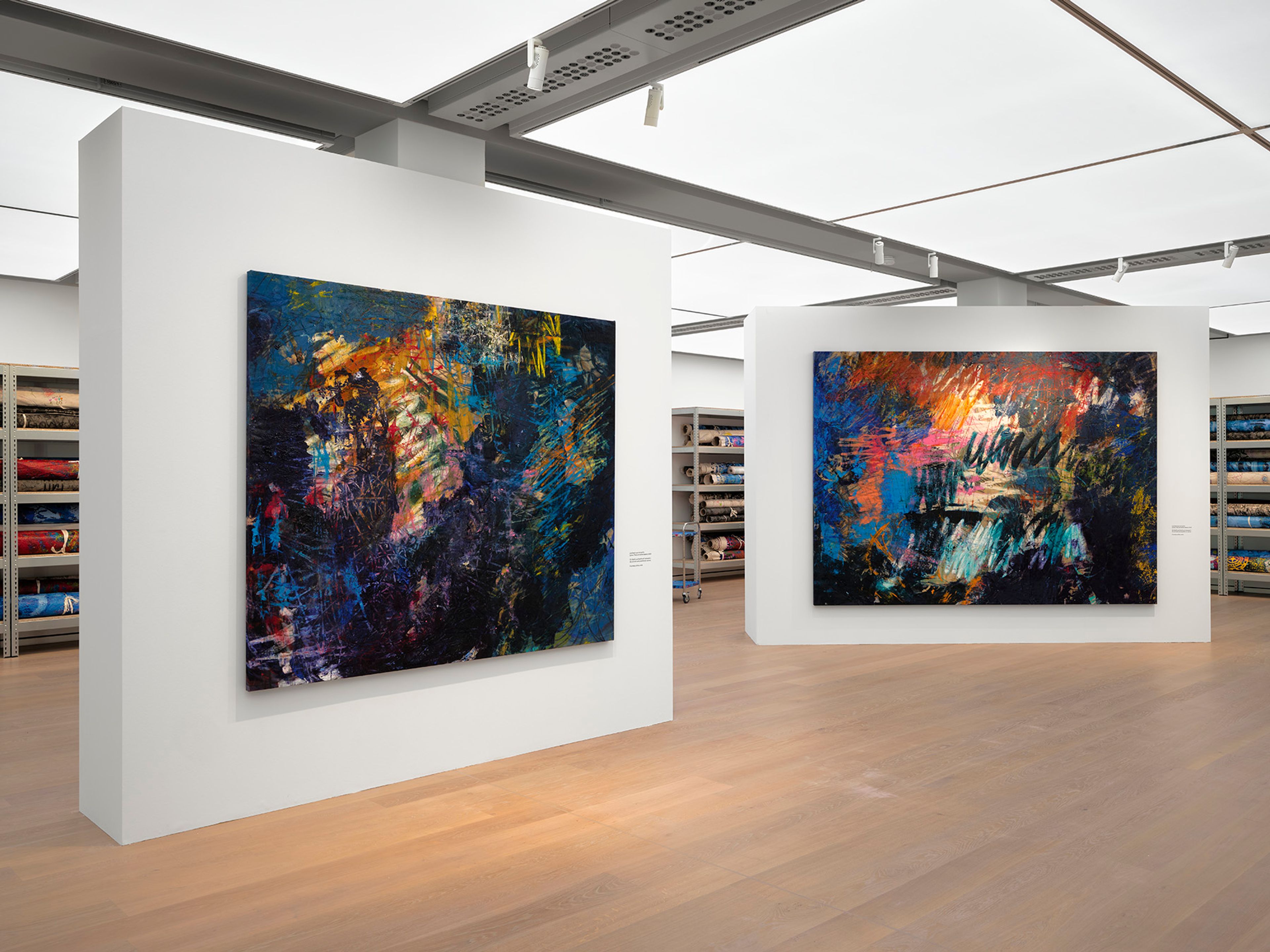 Two large, vibrant abstract paintings with colorful brushstrokes are displayed in a modern gallery setting, flanked by shelves of rolled textiles.