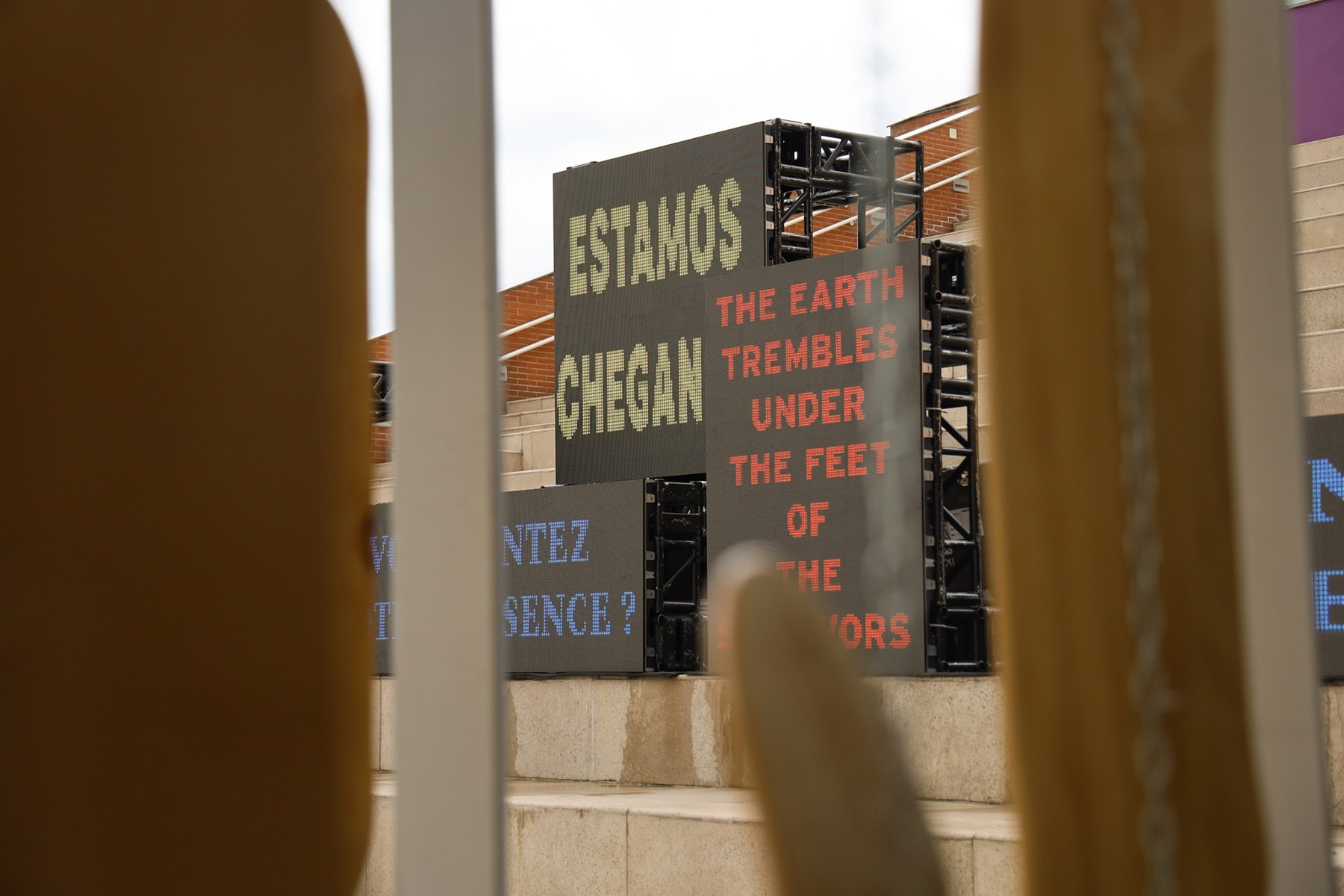 An outdoor installation of dark display boards displaying text: "ESTAMOS CHEGAN" in yellow, and "THE EARTH TREMBLES UNDER THE FEET OF THE SURVIVORS" in red, viewed through a blurry foreground.