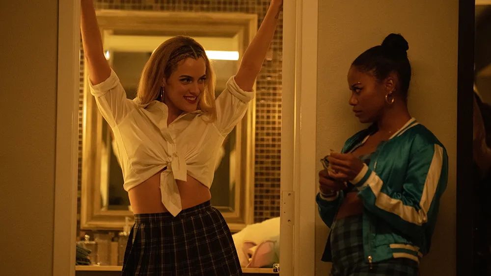 A blonde woman with arms raised and a mischievous smile in a tied white shirt and plaid skirt is watched by a serious dark-haired woman in a teal bomber jacket.