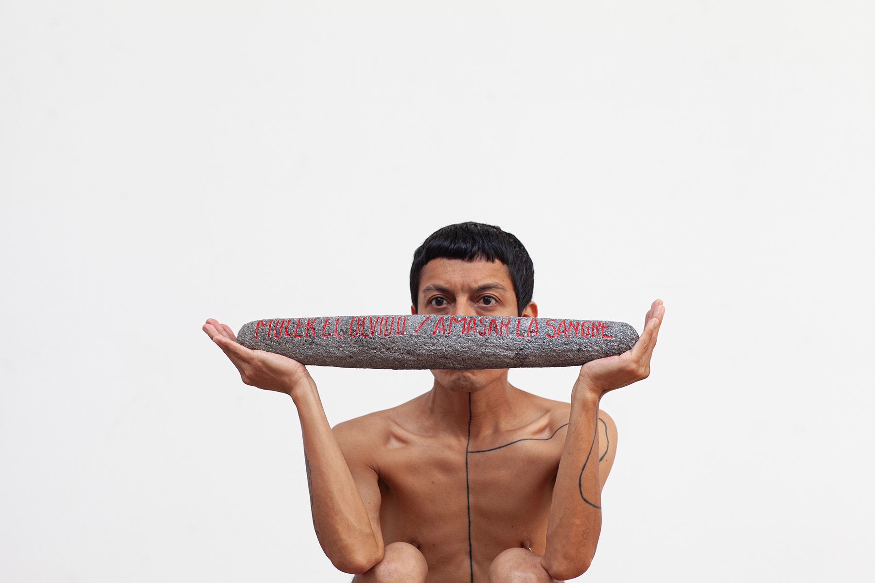 A man with black body markings holds a grey bar with red text "MEZCLAR UN NUEVO AMASAR LA SANGRE" (Mix a new knead the blood) across his mouth, looking intently forward.