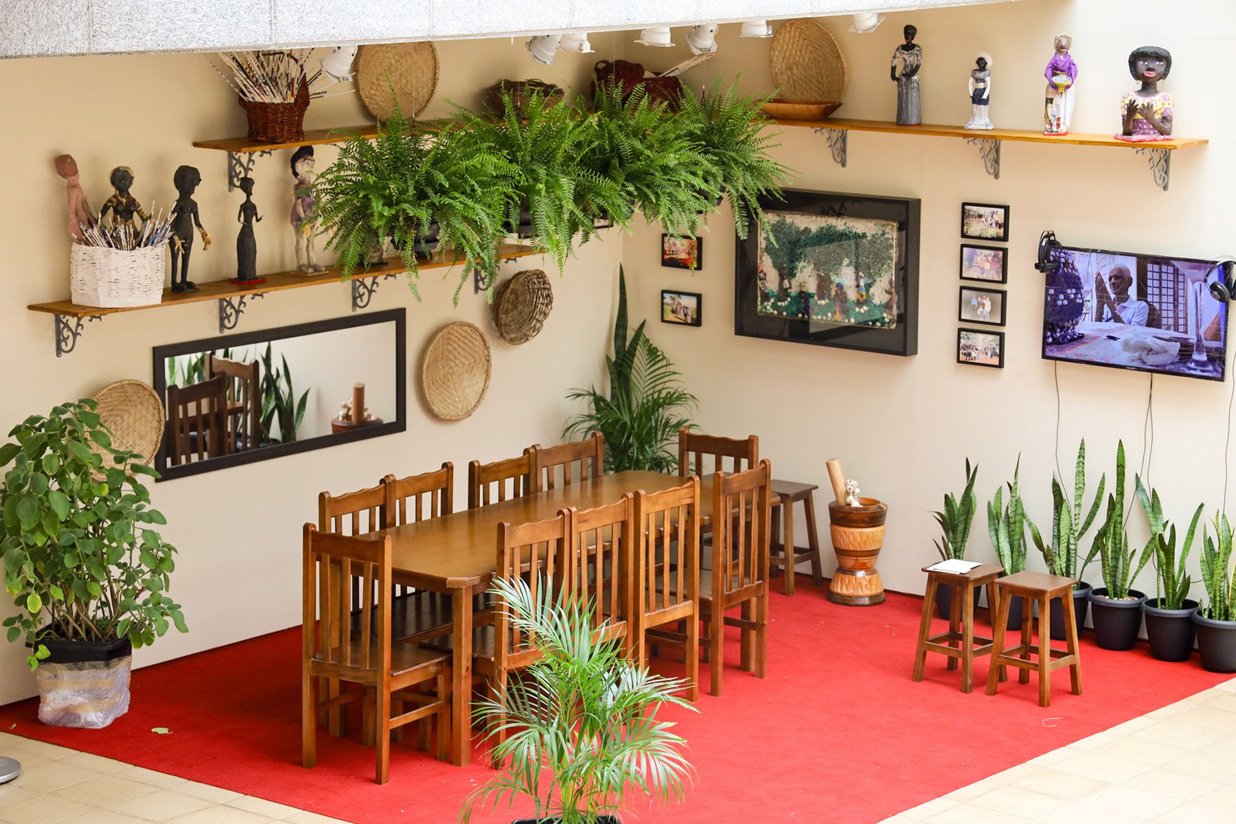 A room with a red floor, a long wooden dining table, shelves displaying plants and cultural figures, and a TV showing a cooking demonstration.