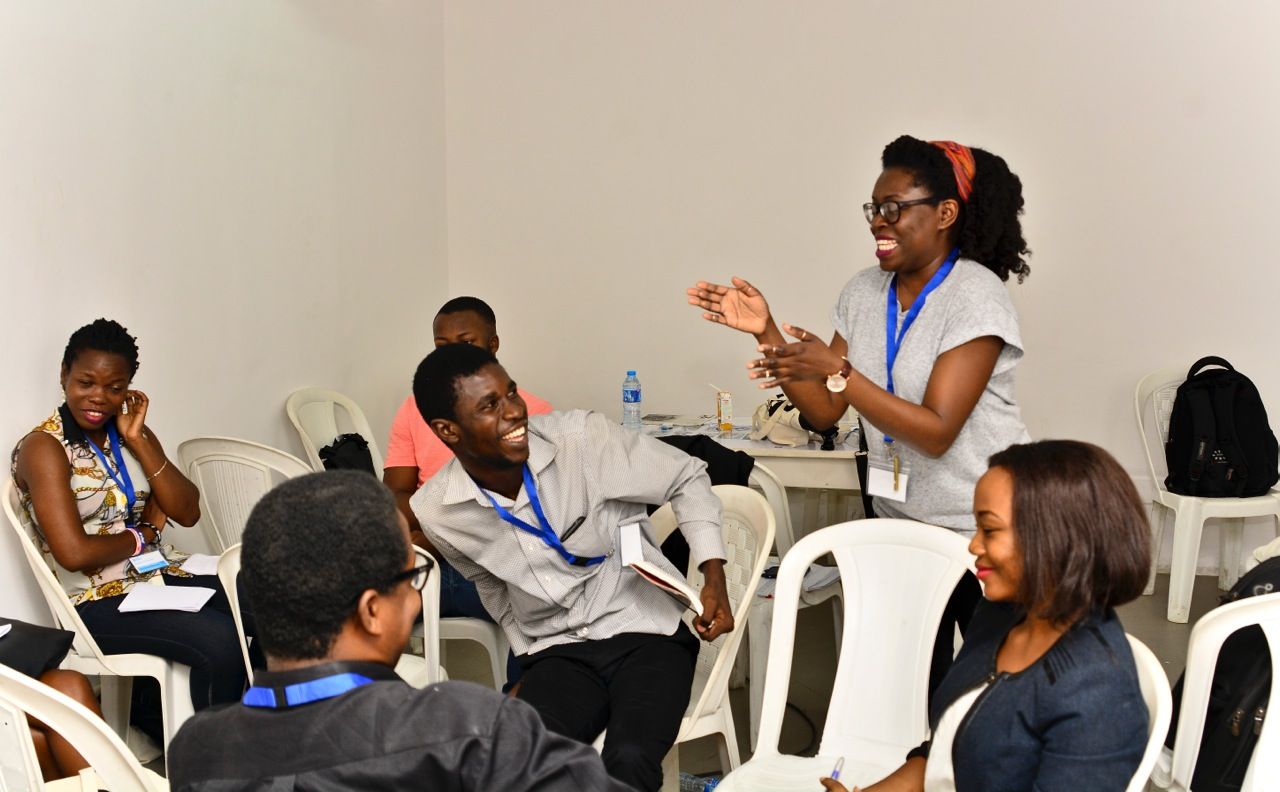 A woman stands laughing and gesturing while speaking to a group of smiling participants at an indoor event.