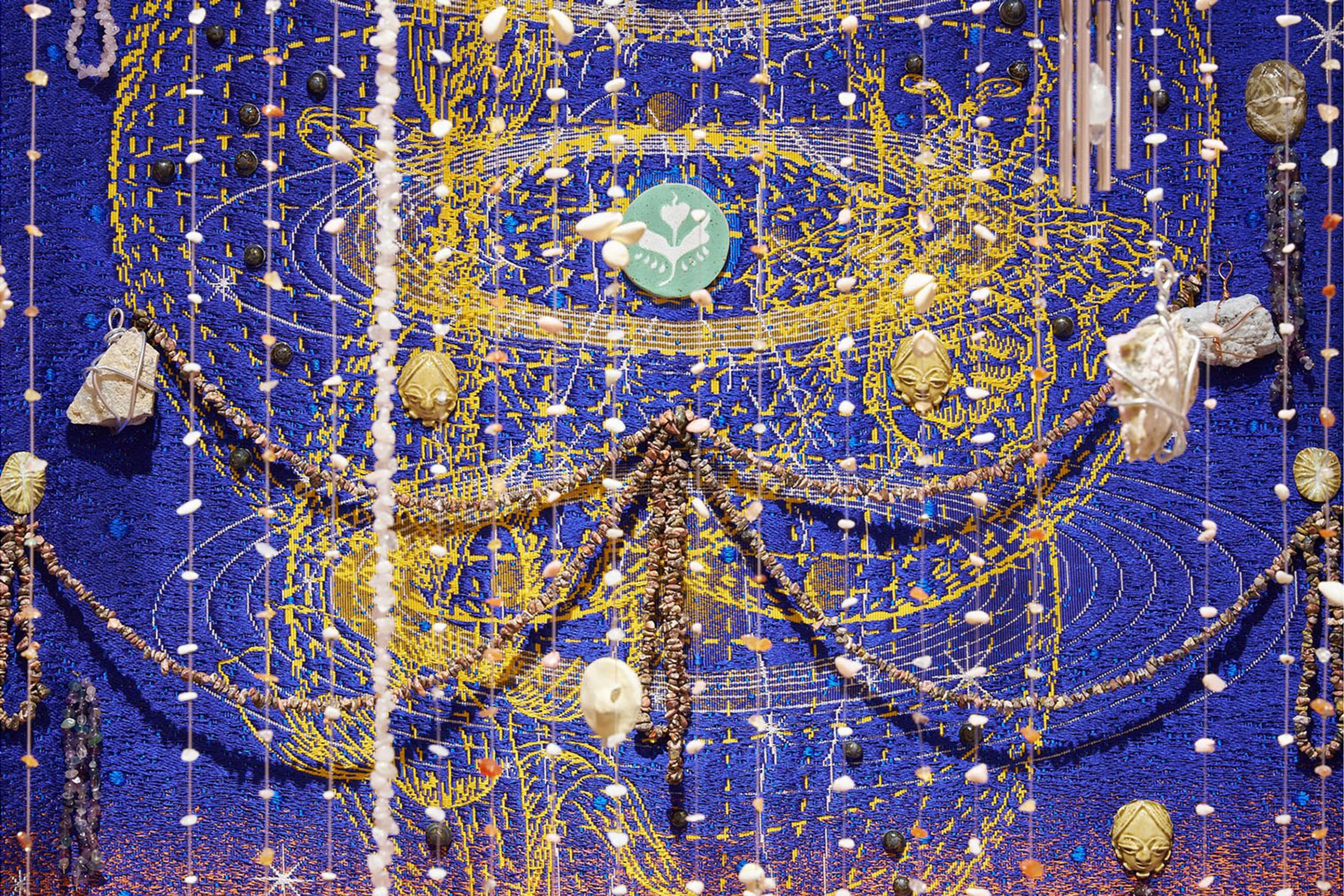 Intricate blue textile art with swirling gold embroidery, adorned with hanging strands of beads, shells, and various natural elements.