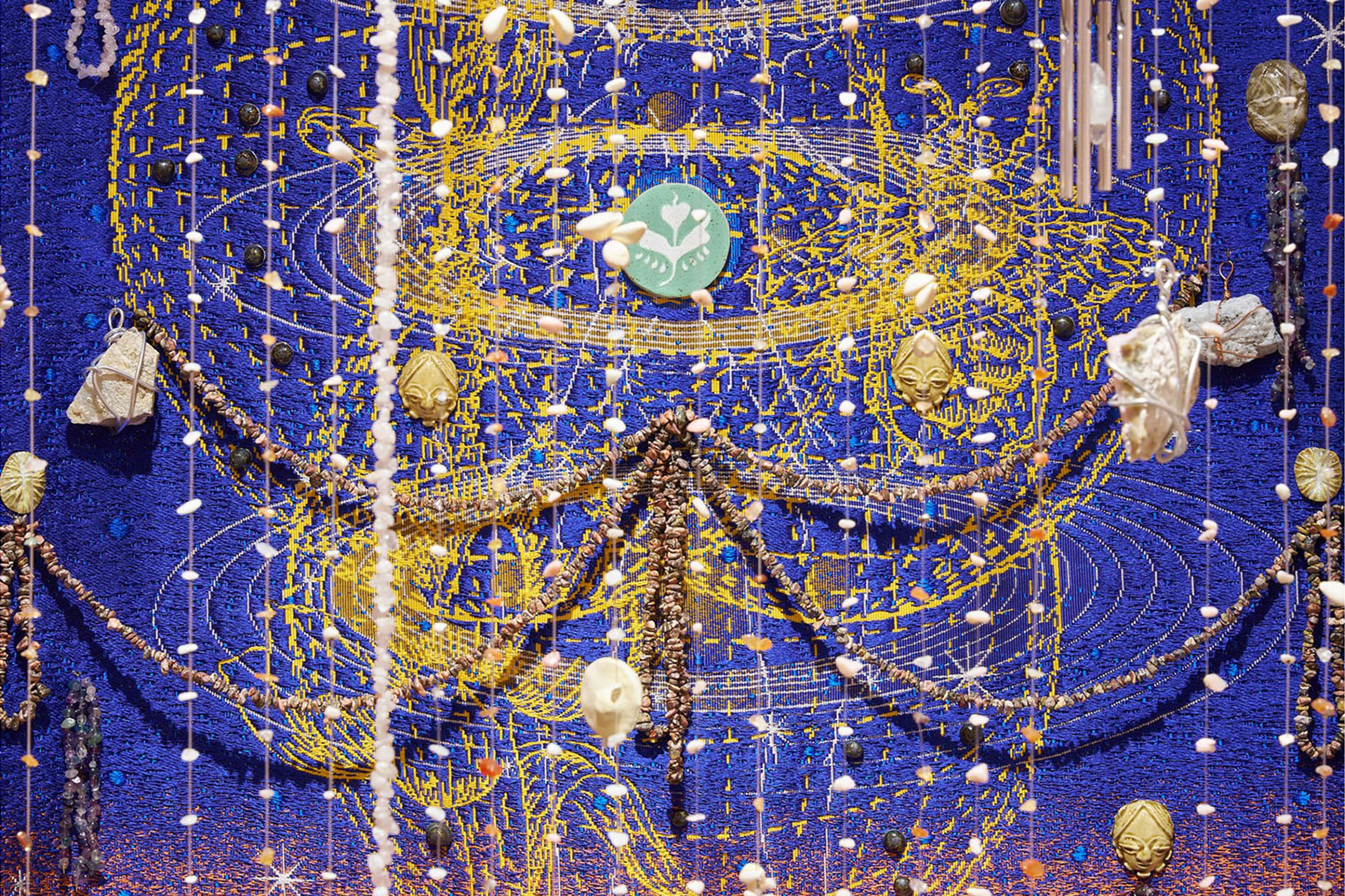 Intricate blue textile art with swirling gold embroidery, adorned with hanging strands of beads, shells, and various natural elements.