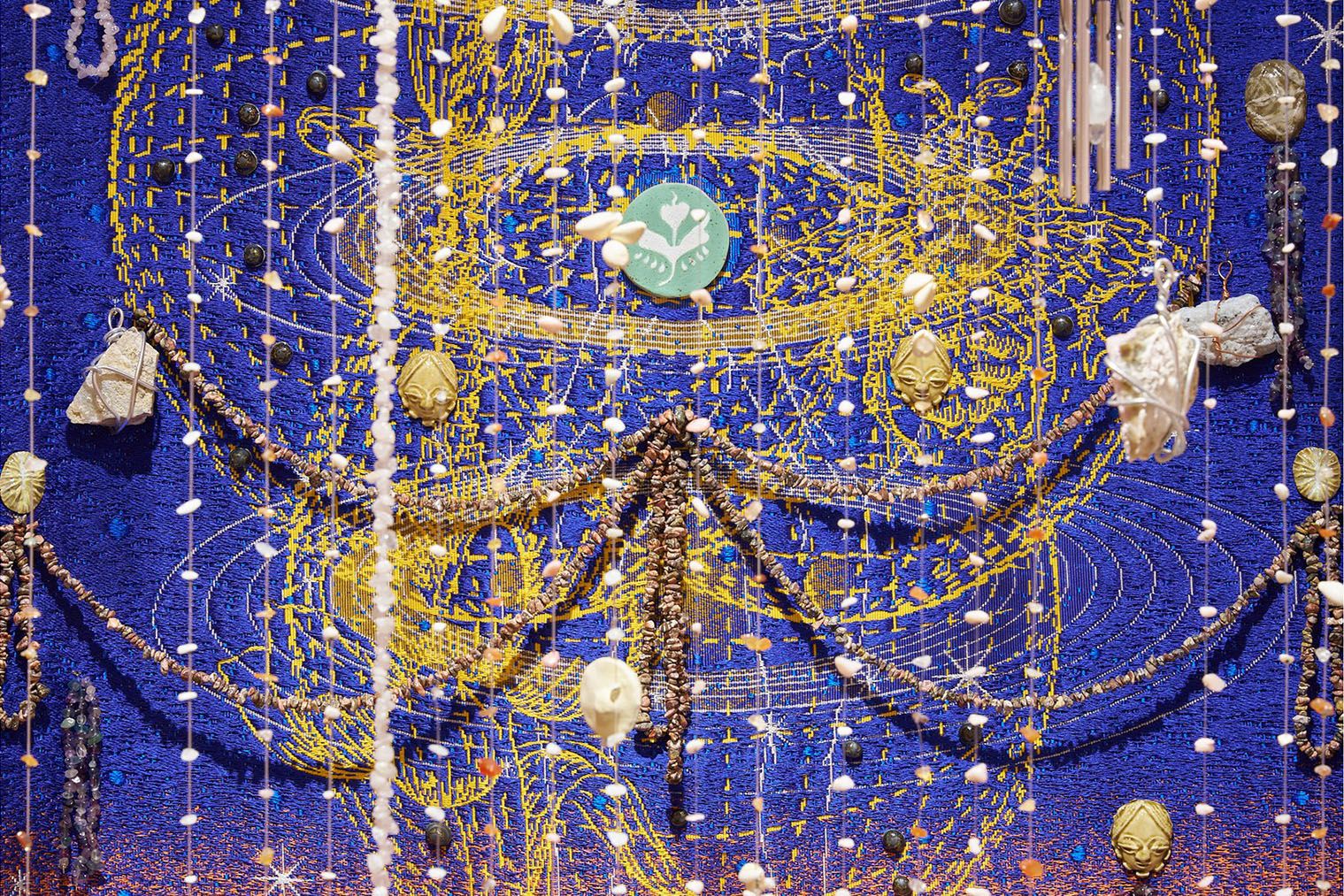 Intricate blue textile art with swirling gold embroidery, adorned with hanging strands of beads, shells, and various natural elements.