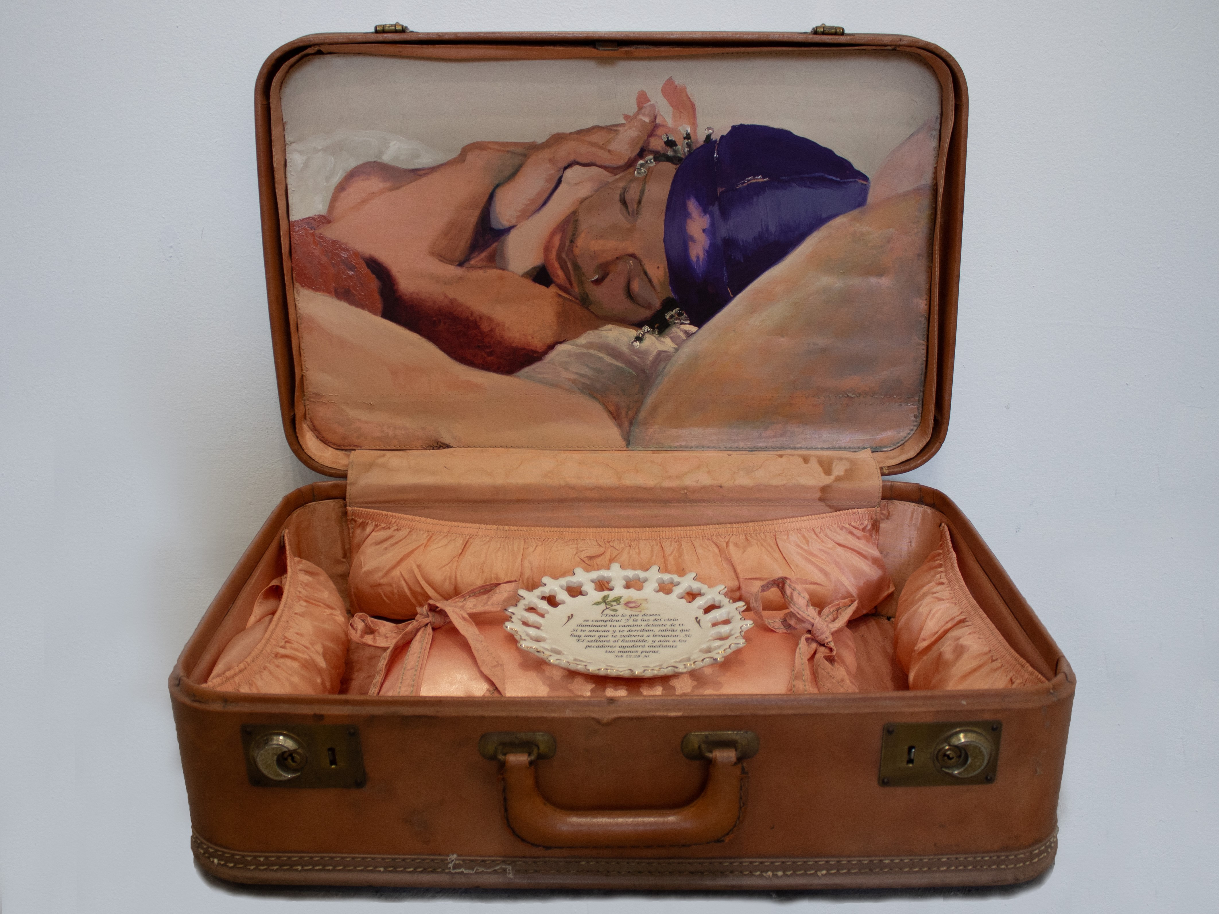 An open vintage suitcase with a painting of a sleeping person in its lid, and a peach-lined interior holding a decorative white plate.