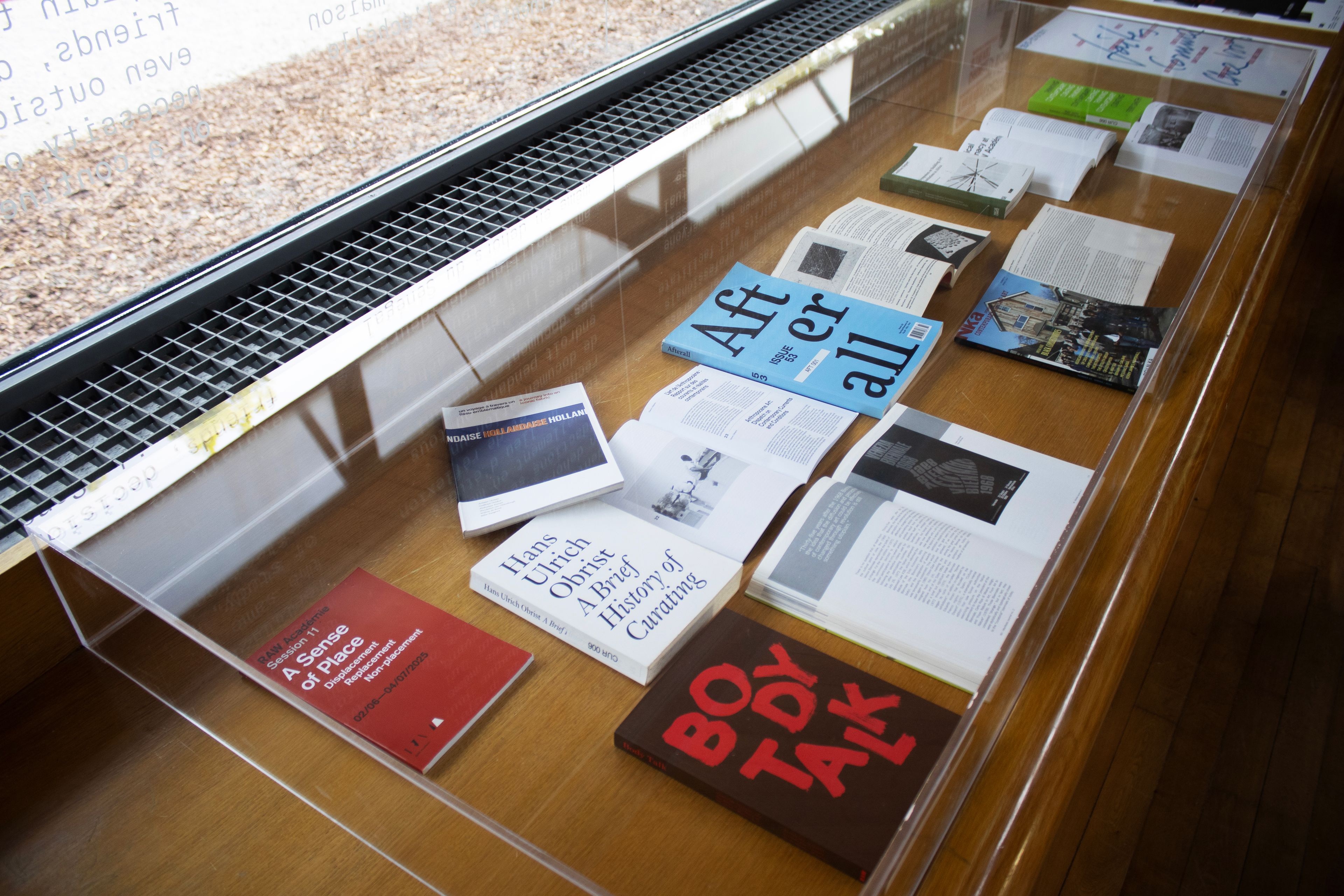 A clear acrylic display case holds various art and culture books and magazines, some open, on a wooden table.
