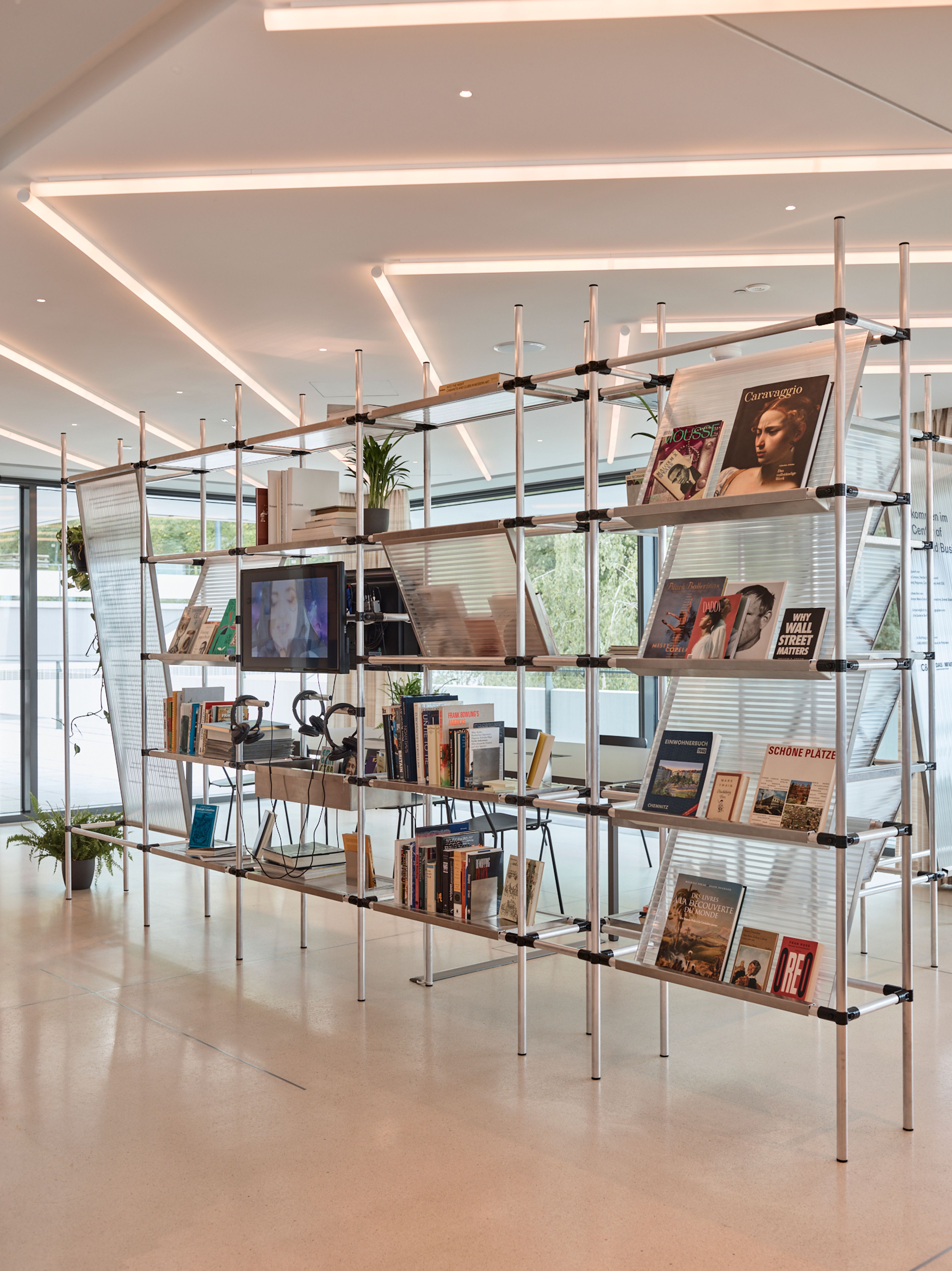 A modern, multi-level shelving unit filled with books, magazines, a TV, headphones, and plants, dividing a bright, contemporary space.