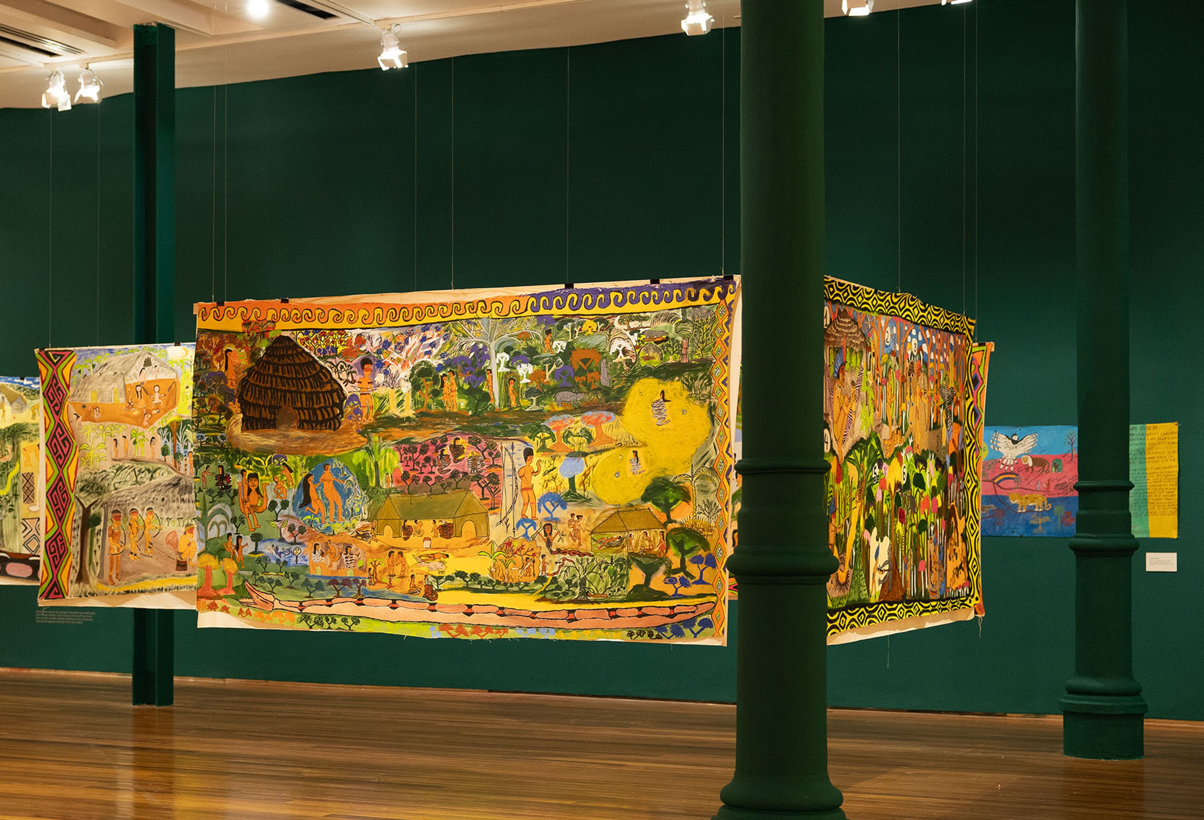Vibrant textile art depicting narrative scenes hangs in a dark green gallery.