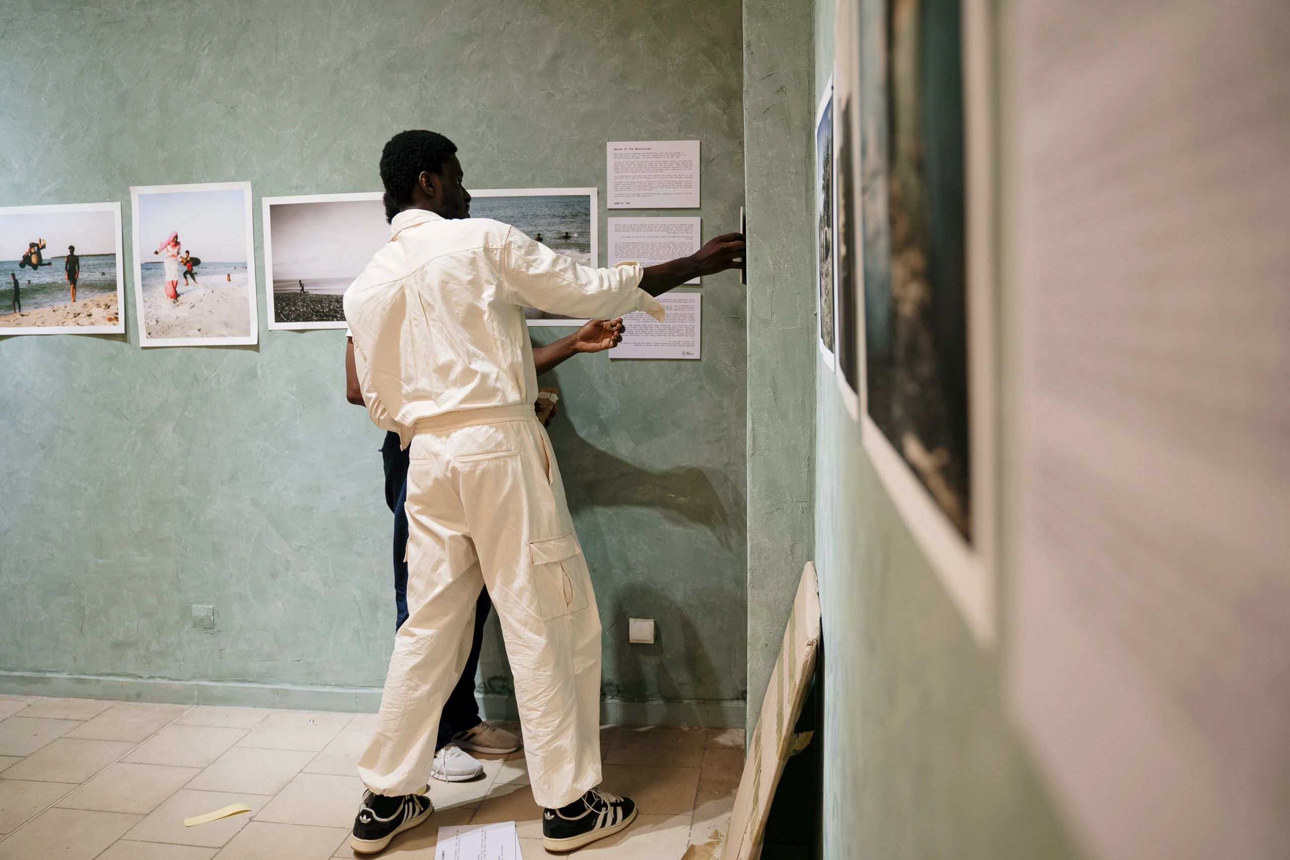 A person in white overalls hangs framed artwork and text on a light green gallery wall.
