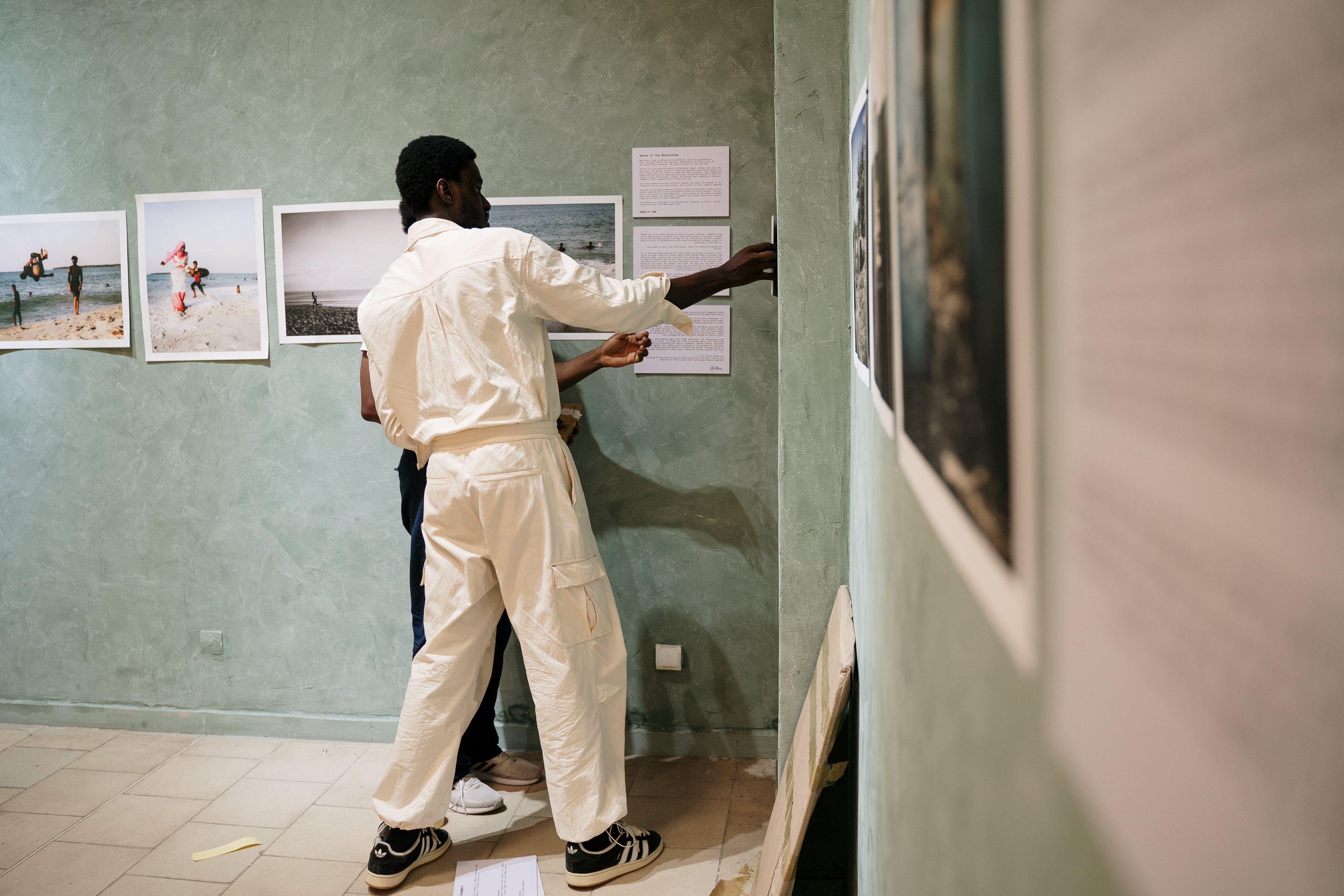 A person in white overalls hangs framed artwork and text on a light green gallery wall.
