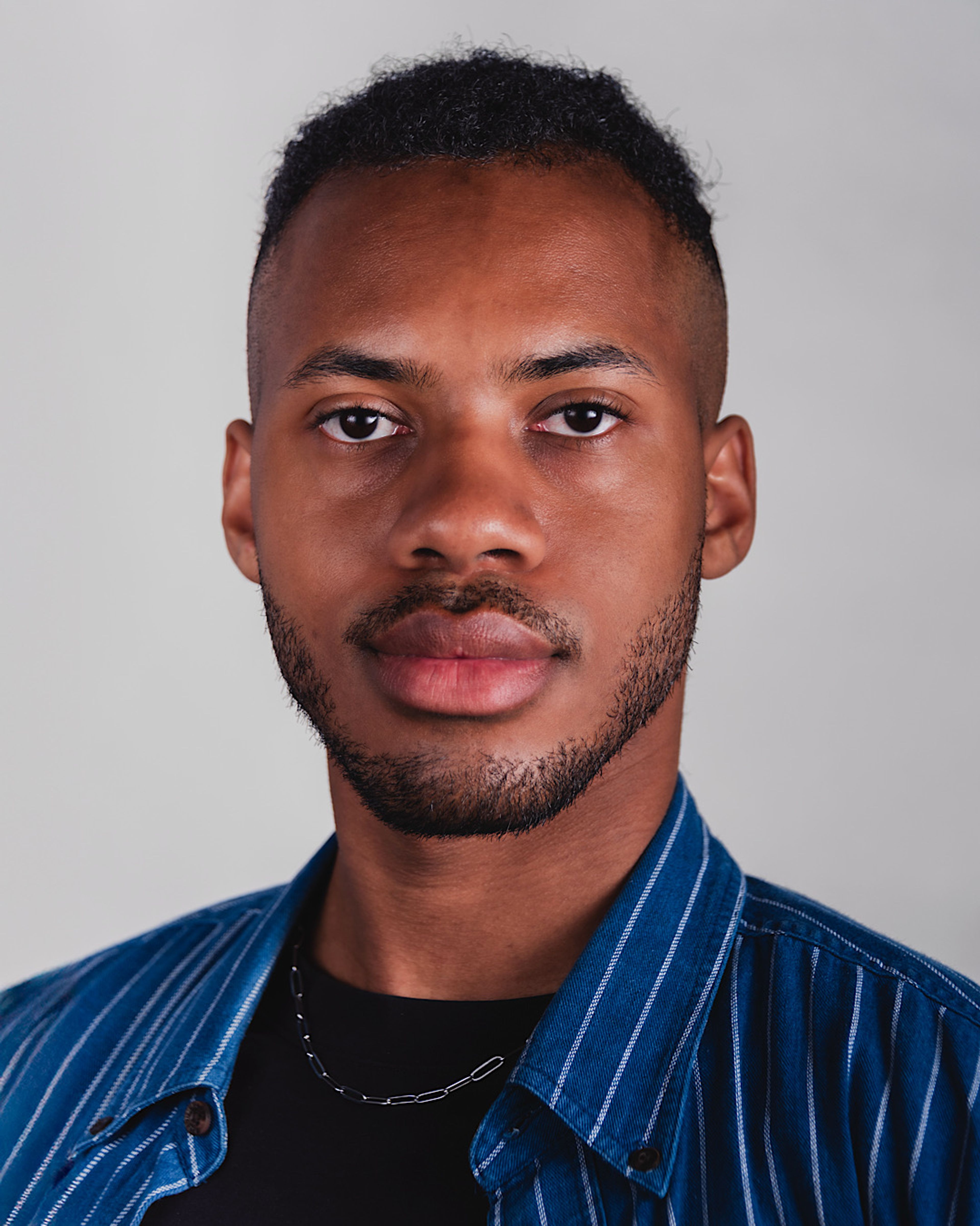 Young Black man with short hair and a light beard, wearing a blue striped shirt and a silver chain, looking at the viewer.