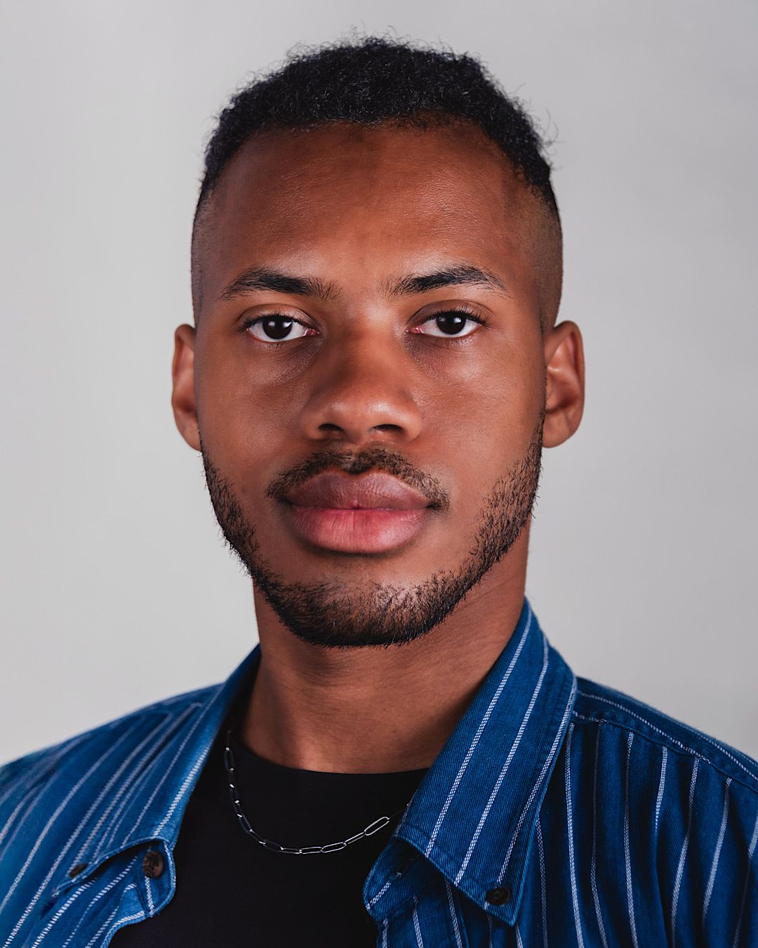 Young Black man with short hair and a light beard, wearing a blue striped shirt and a silver chain, looking at the viewer.