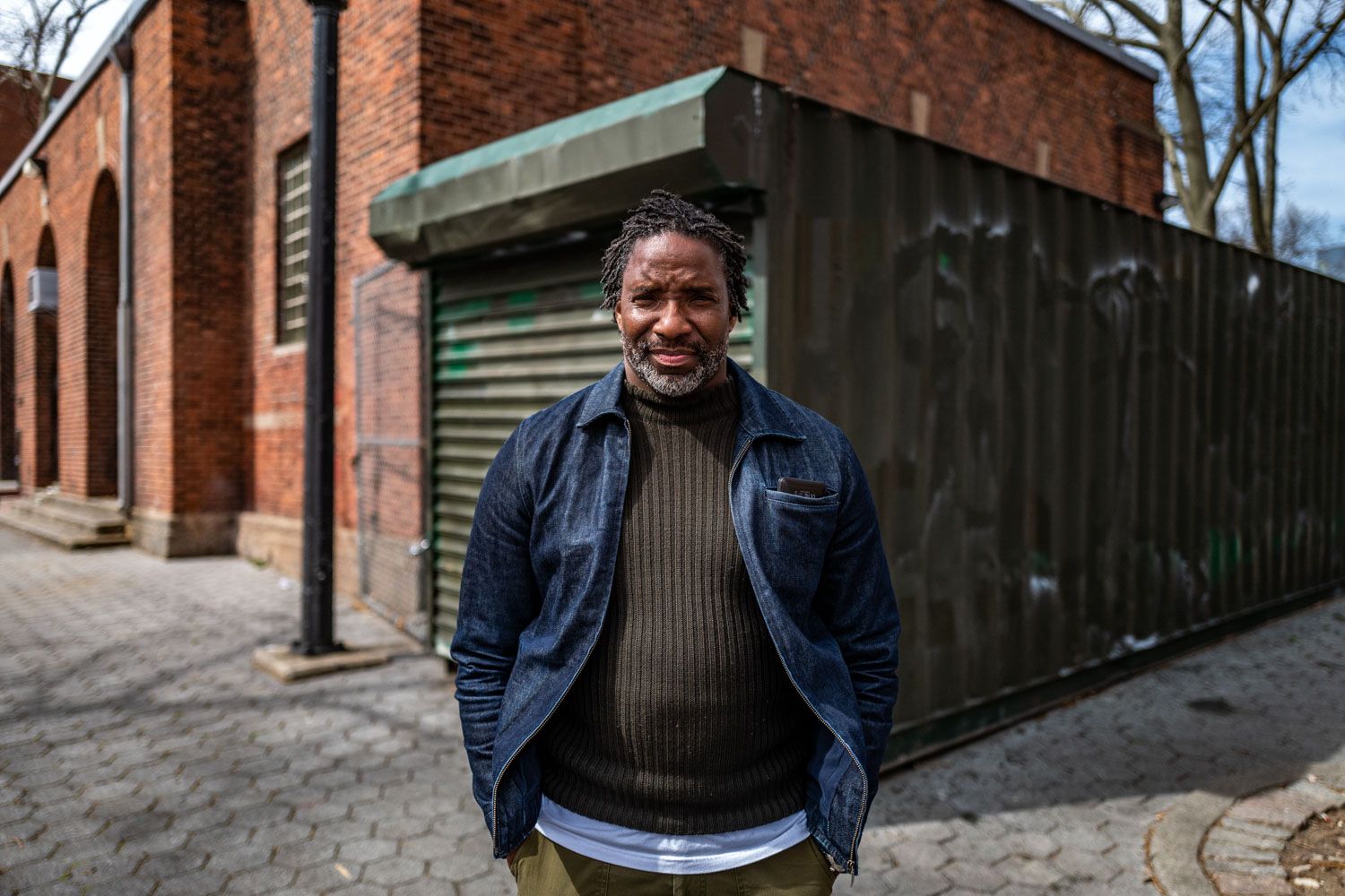 A man with dreadlocks and a beard stands outdoors, looking at the camera, with a brick building and dark metal structure behind him.