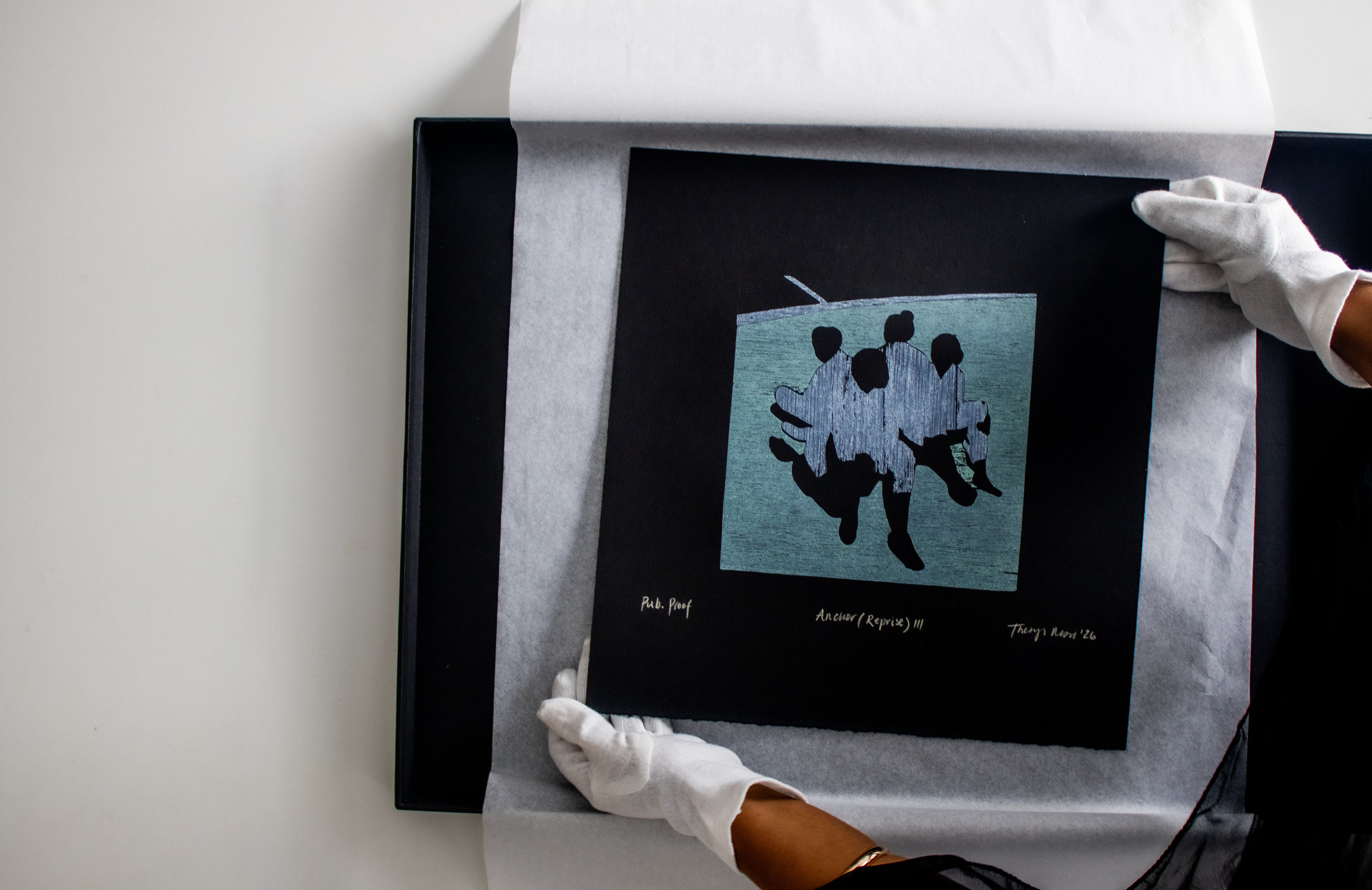 A person in white gloves places a black print depicting four abstract blue and black figures into a black frame.