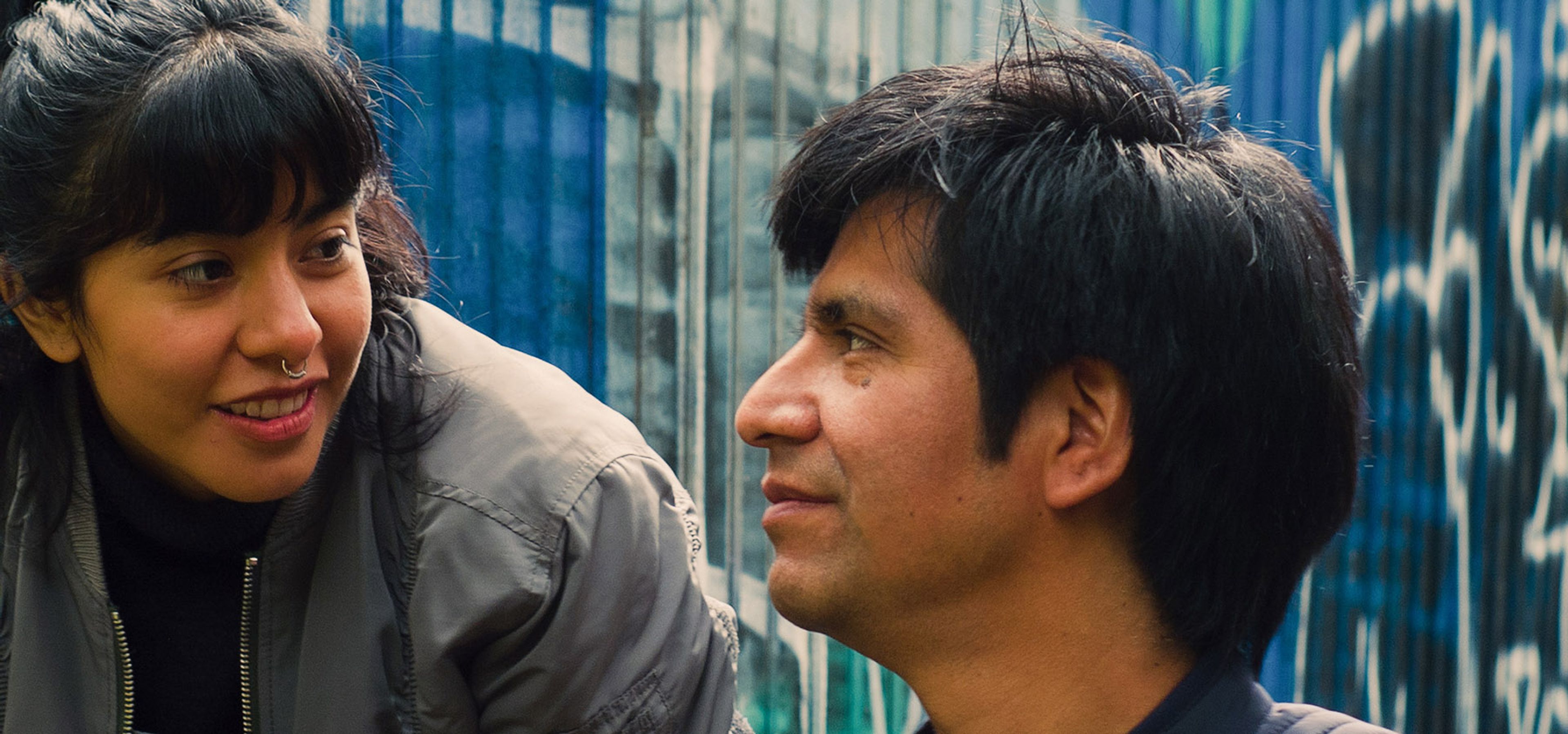 A smiling woman with a nose ring looks toward a man in profile, against a graffiti wall.